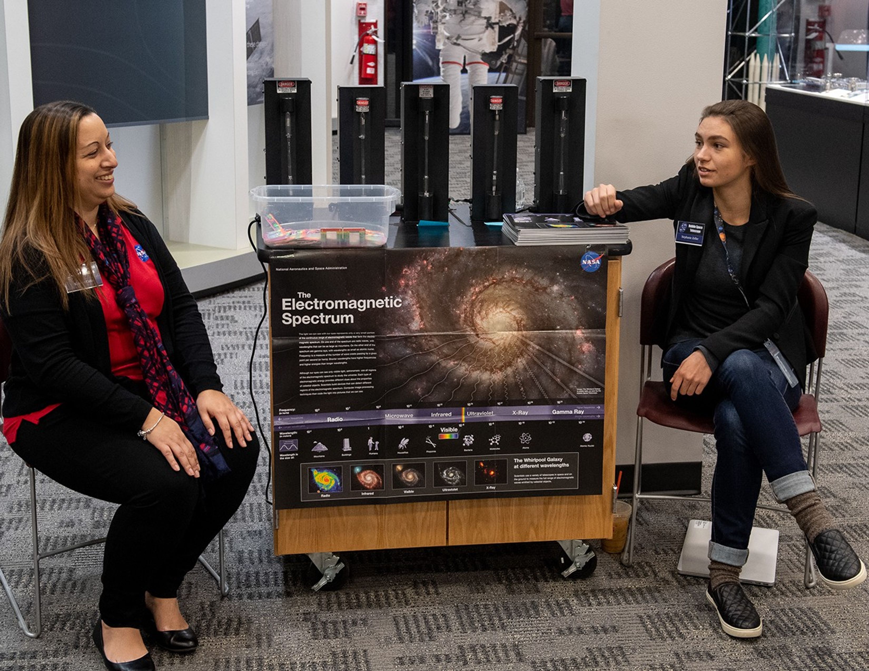 Colon (left) and Zeller (right) sit talking with an activity cart between them; it has a poster about the electromagnetic spectrum attached to the front, with a picture of a galaxy on it. Five black devices with glass light tubes, a plastic container, and a pile of handouts sit on the cart.