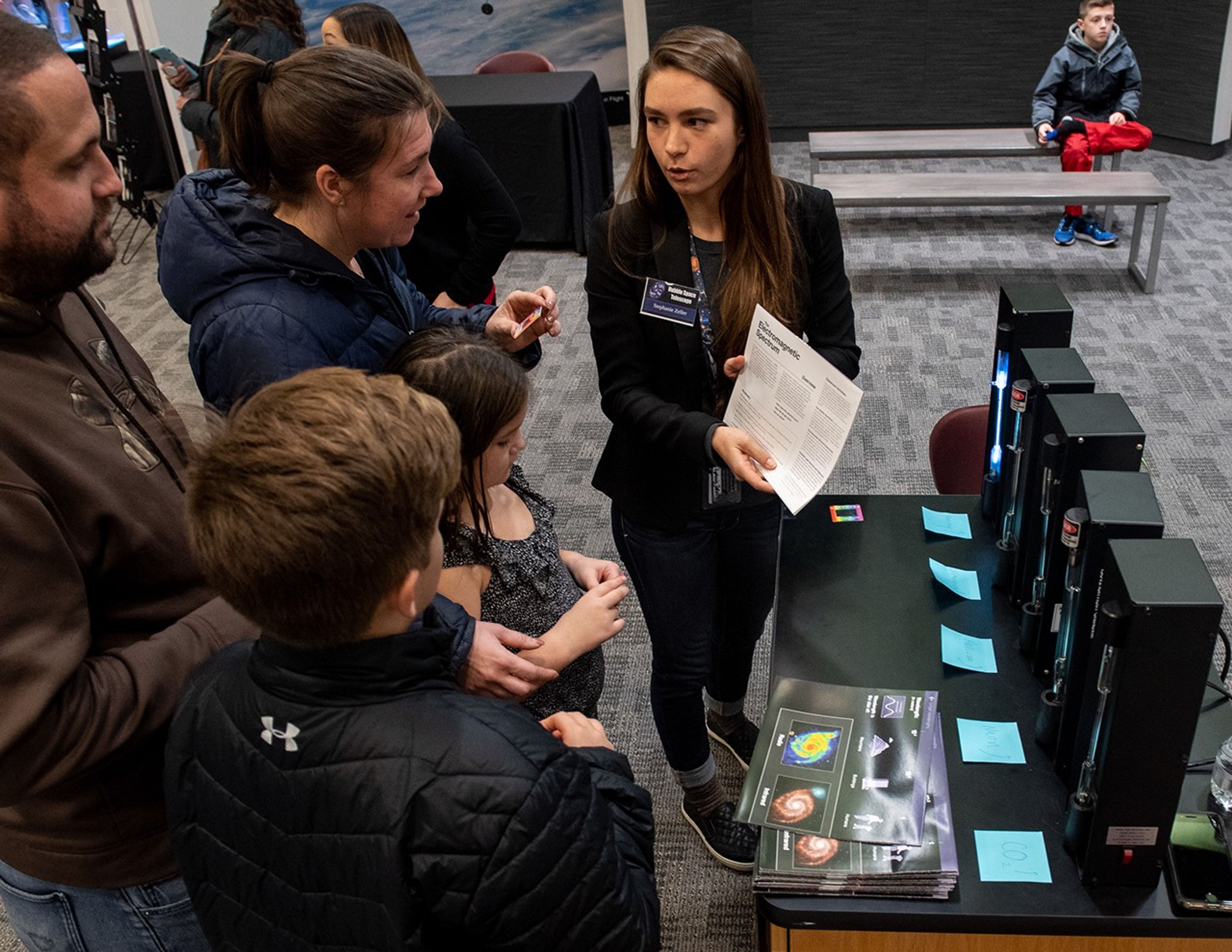 Stephanie Zeller, right, shows a family of four, left, the back of a handout while demonstrating the spectrum tube activity on the right side of the picture. More visitors are visible in the background.