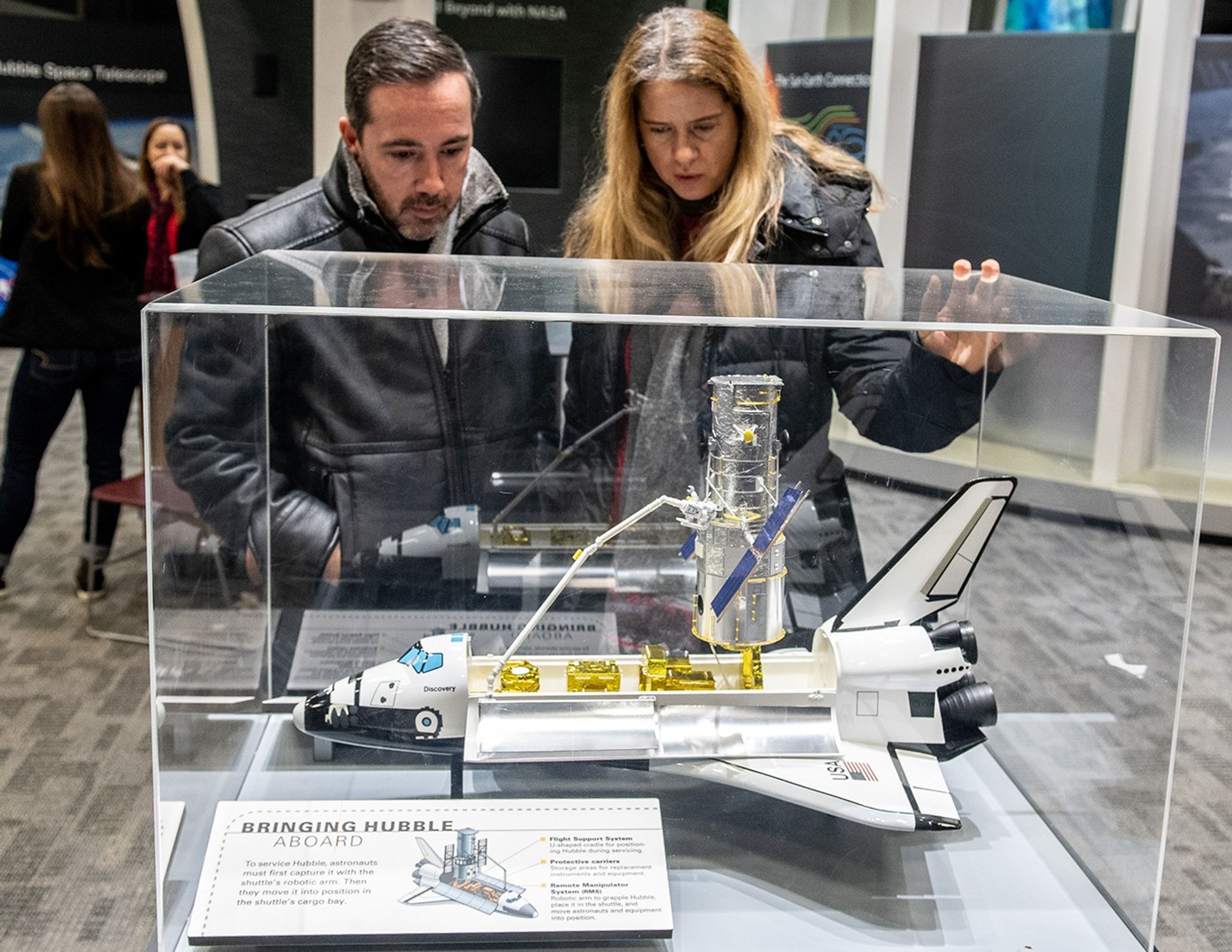 Two visitors, a man and a woman, examine the model of the Discovery space shuttle with a model of Hubble in its bay, contained in a glass case between them and the camera. Two NASA employees are visible in the background on the left.
