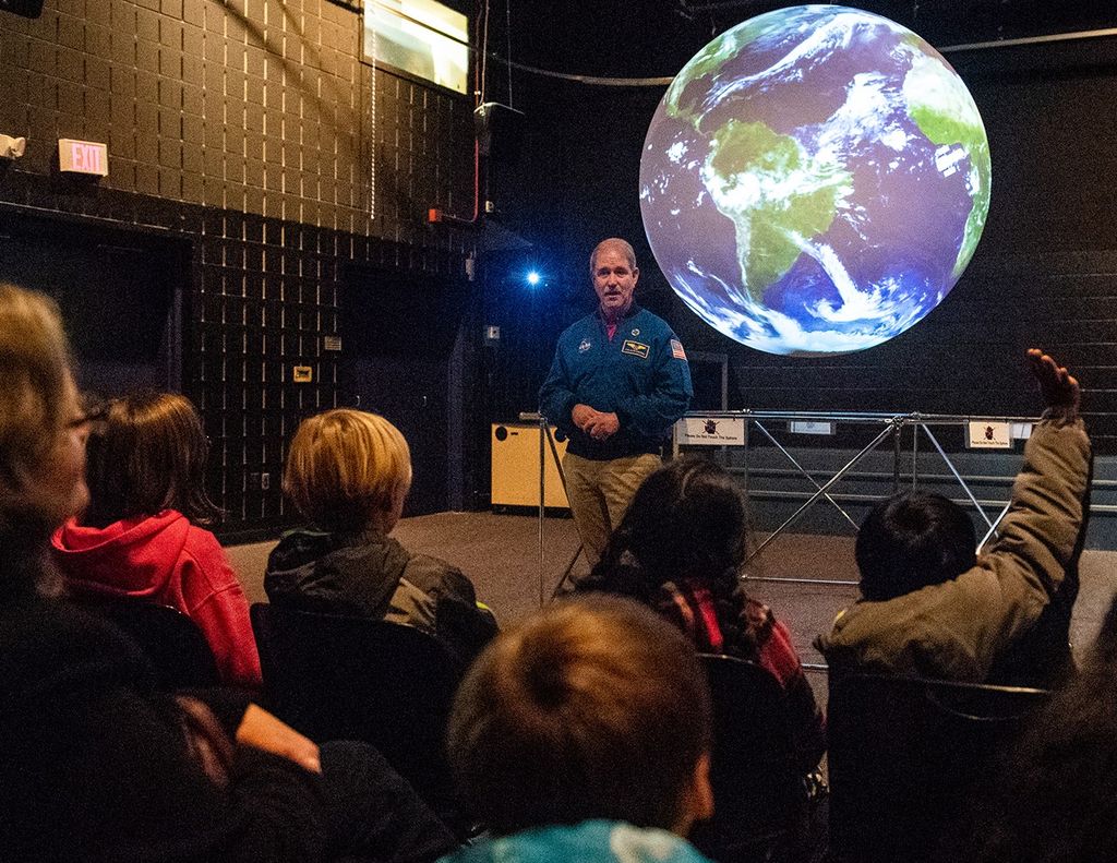 John Grunsfeld stands in front of the Science on a Sphere display, on which the display looks like a "globe" of Earth. A group of students and chaperones sit in the foreground, facing him; the student on the far-right has a hand raised.