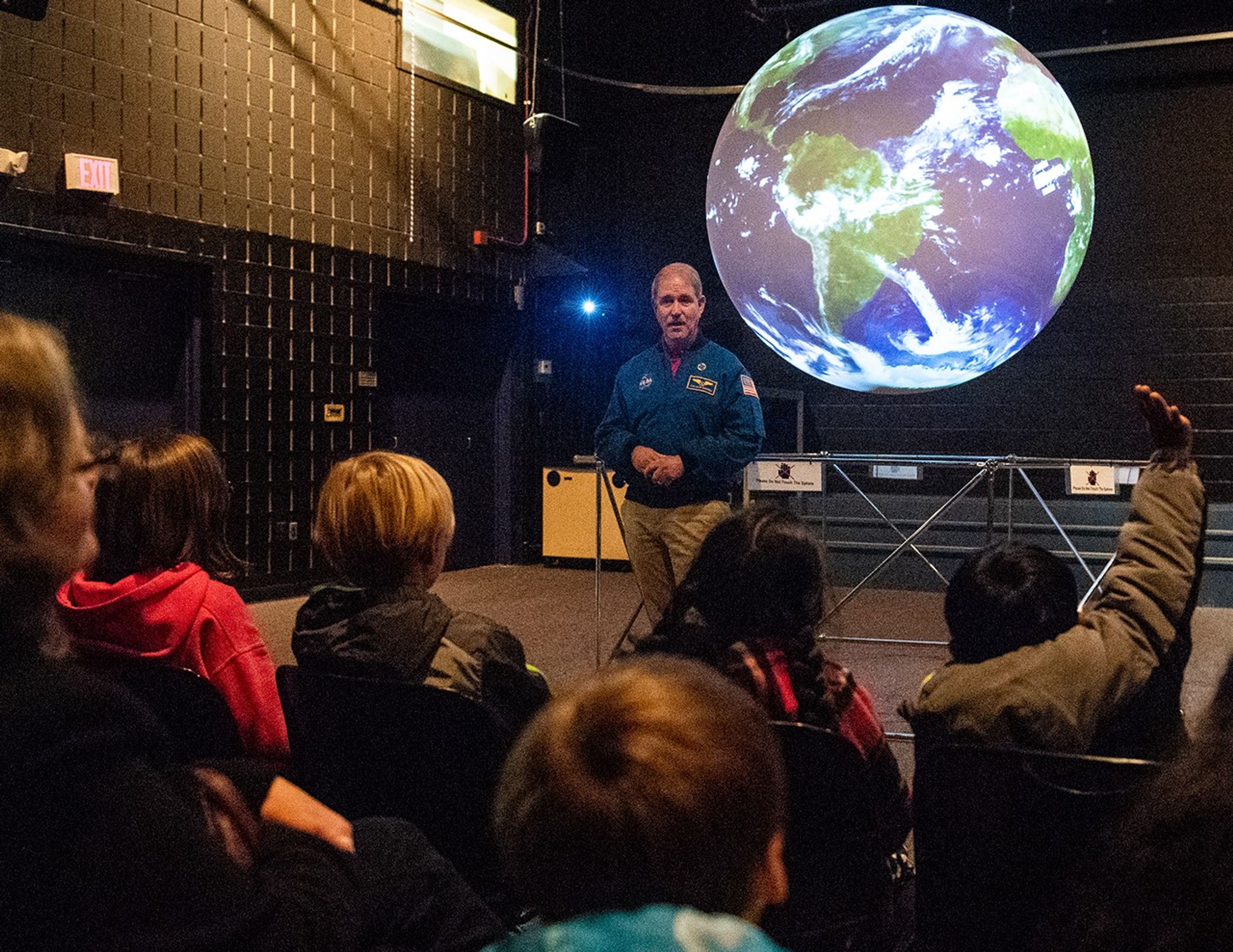John Grunsfeld stands in front of the Science on a Sphere display, on which the display looks like a "globe" of Earth. A group of students and chaperones sit in the foreground, facing him; the student on the far-right has a hand raised.