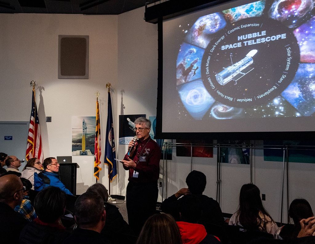 Jim Jeletic, microphone in hand, speaks to a listening audience, with a slide featuring the Hubble Space Telescope projected onto the screen behind him.