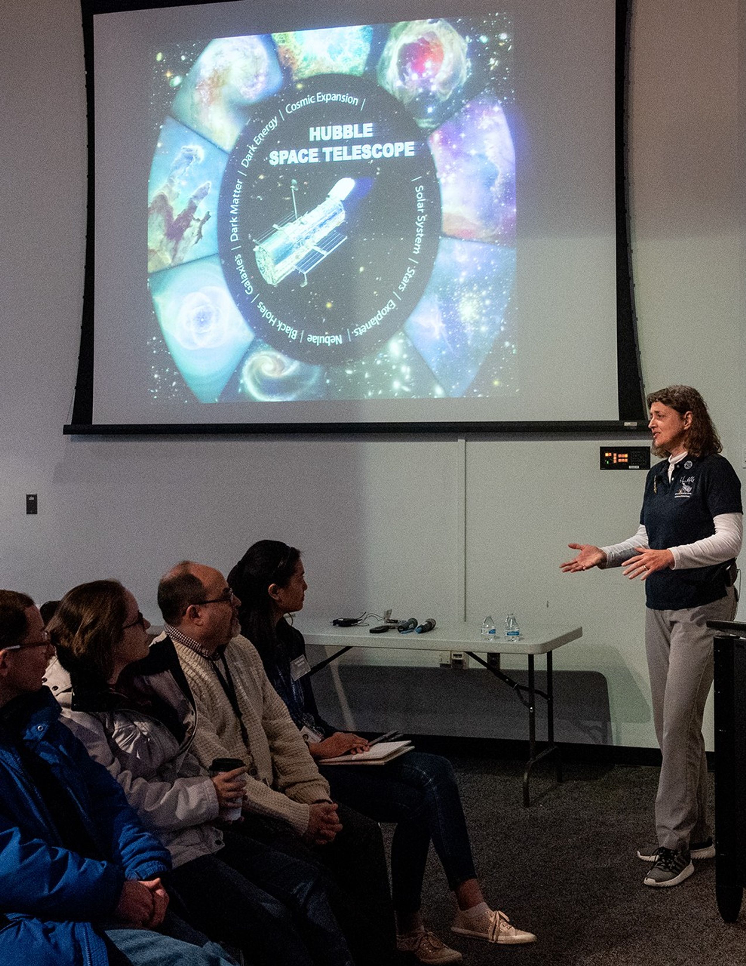 Jennifer Wiseman, right, speaks to a listening audience, left, with a "Hubble Space Telescope" intro slide projected onto the screen above them.