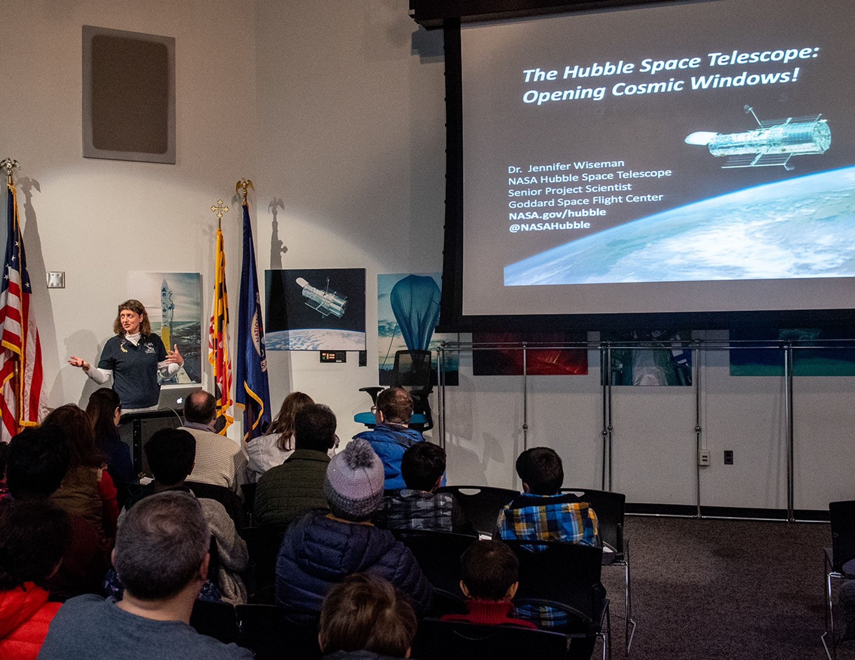 Jennifer Wiseman, left, speaks to a listening audience, with an intro slide projected onto the screen in the upper right.