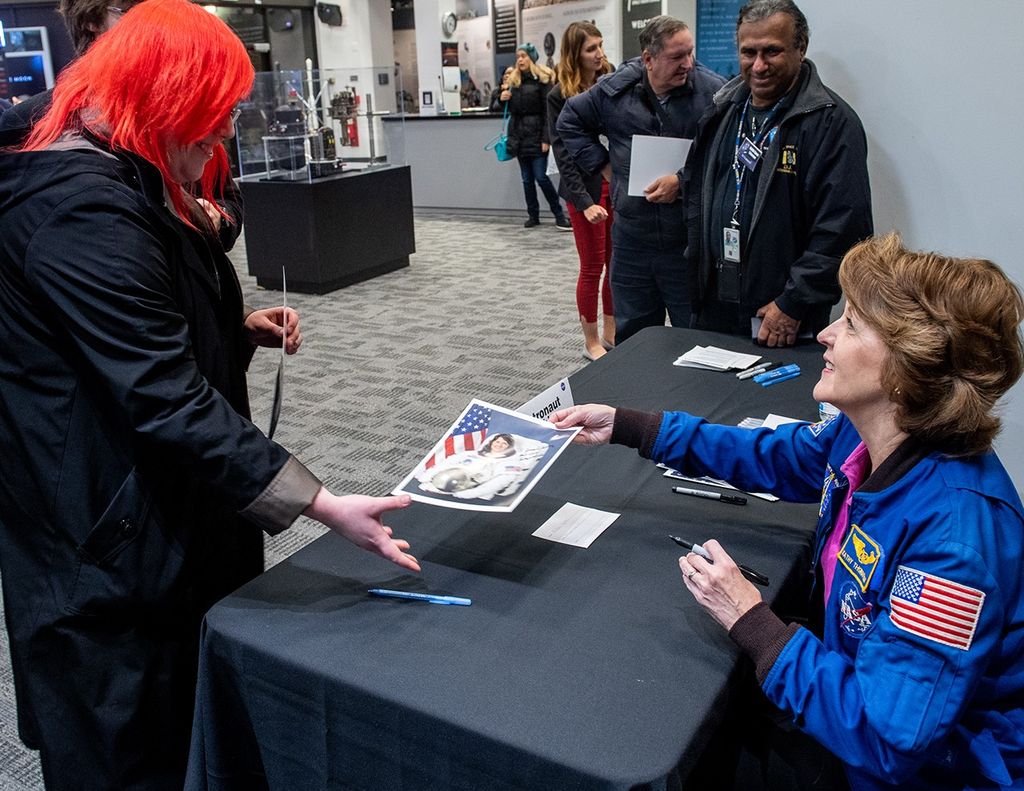 Kathy Thornton (sitting behind a table on the right) signs one of her astronaut pictures for a visitor (left) with several other visitors and staff visible in the background.