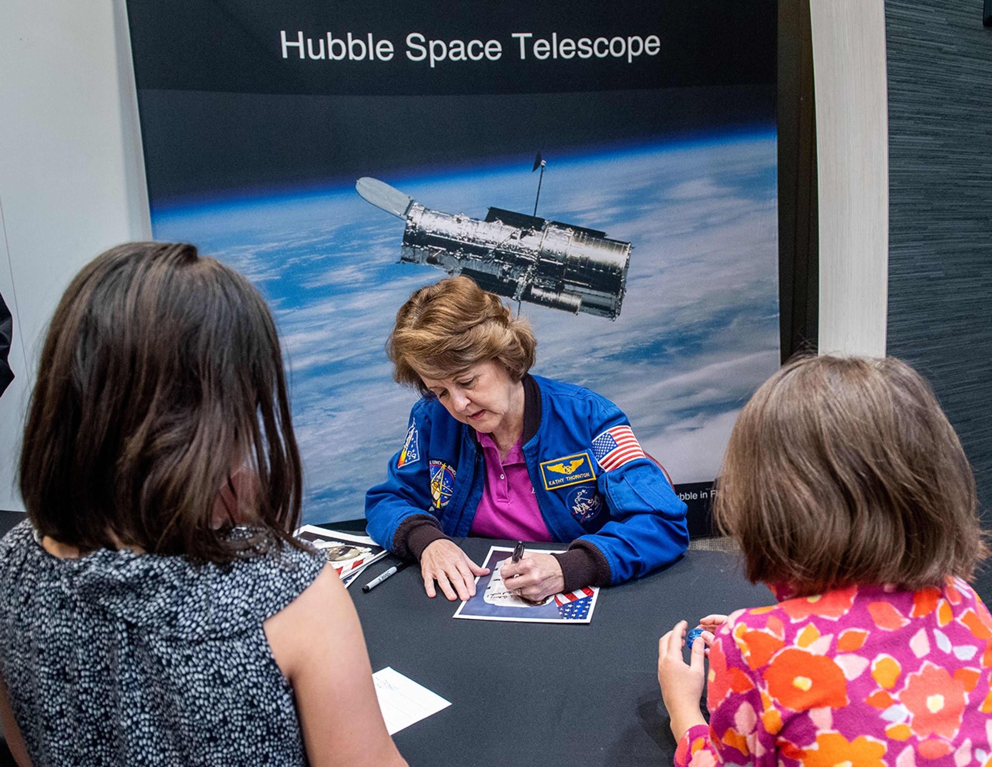Kathy Thornton, sitting behind the table with an image of the Hubble Space Telescope behind her, signs an astronaut for two young visitors in the foreground.