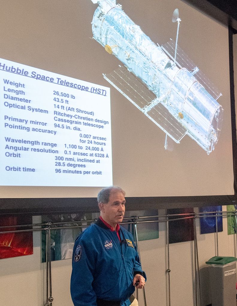 John Grunsfeld speaks, with an image of Hubble projected onto the screen behind him.