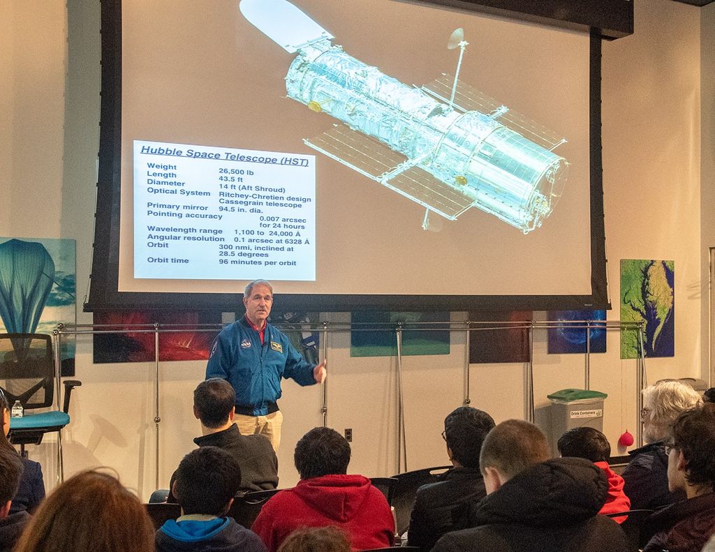 John Grunsfeld speaks to a listening audience, with an image of Hubble projected onto the screen above him.