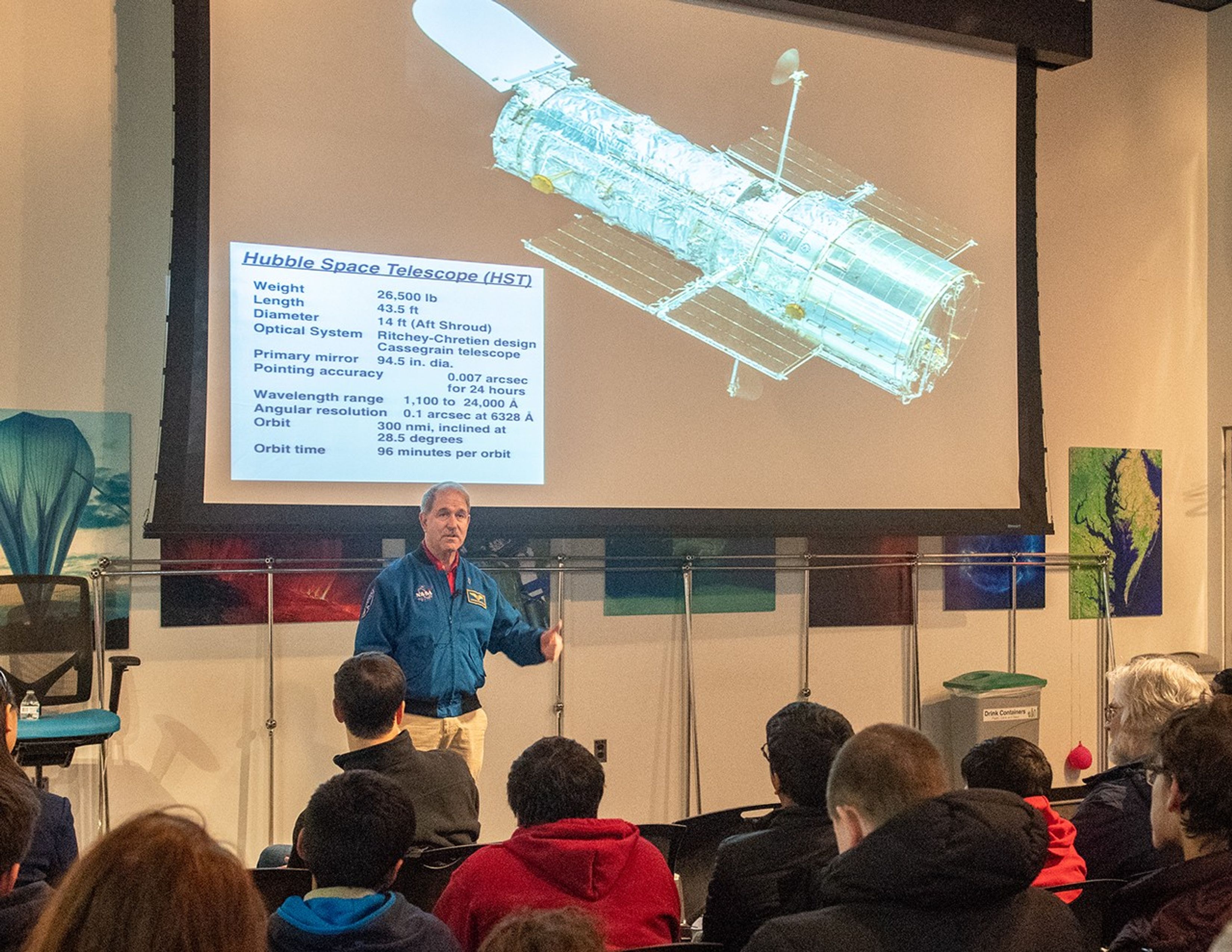 John Grunsfeld speaks to a listening audience, with an image of Hubble projected onto the screen above him.