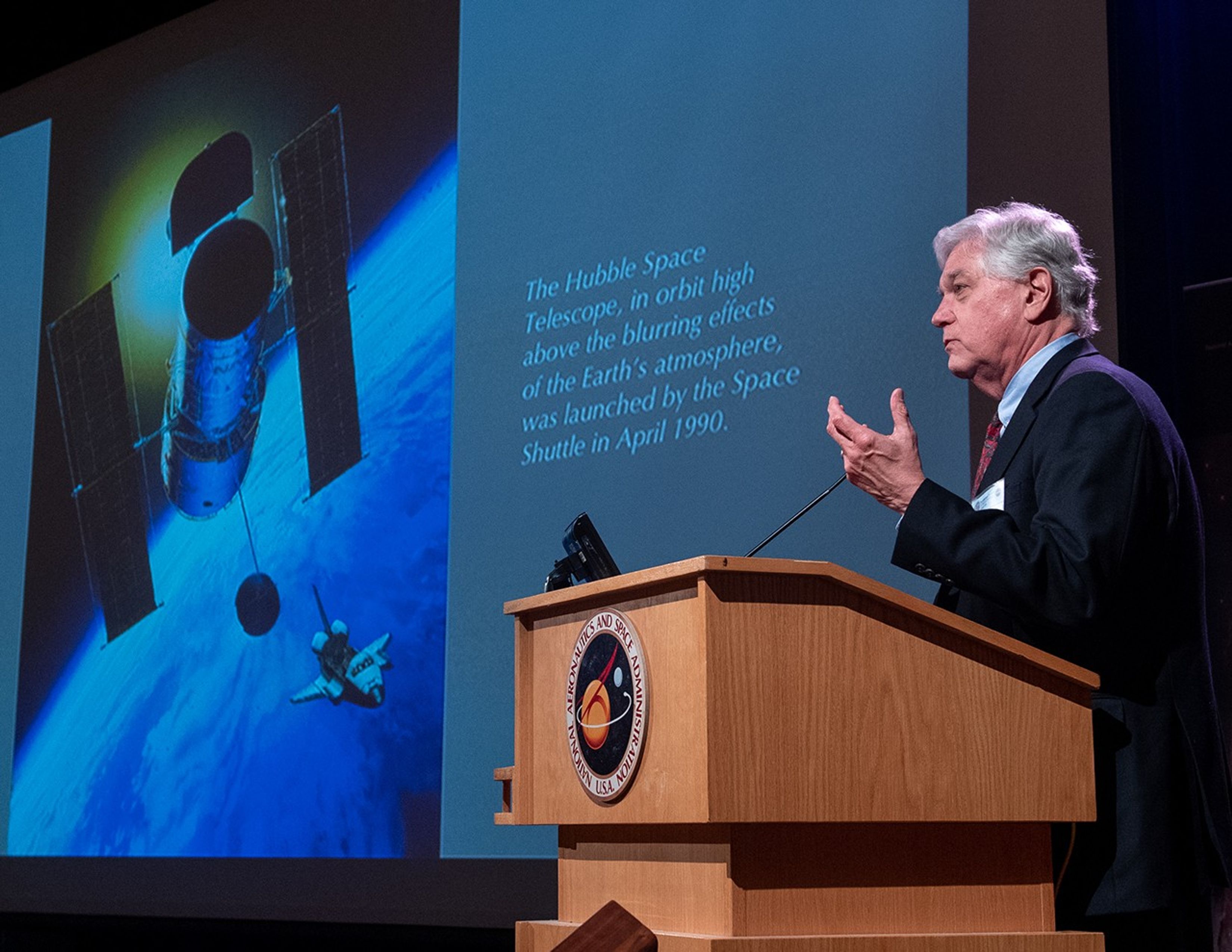 John Trauger stands at the NASA podium addressing the audience, with the slide show visible in the background.