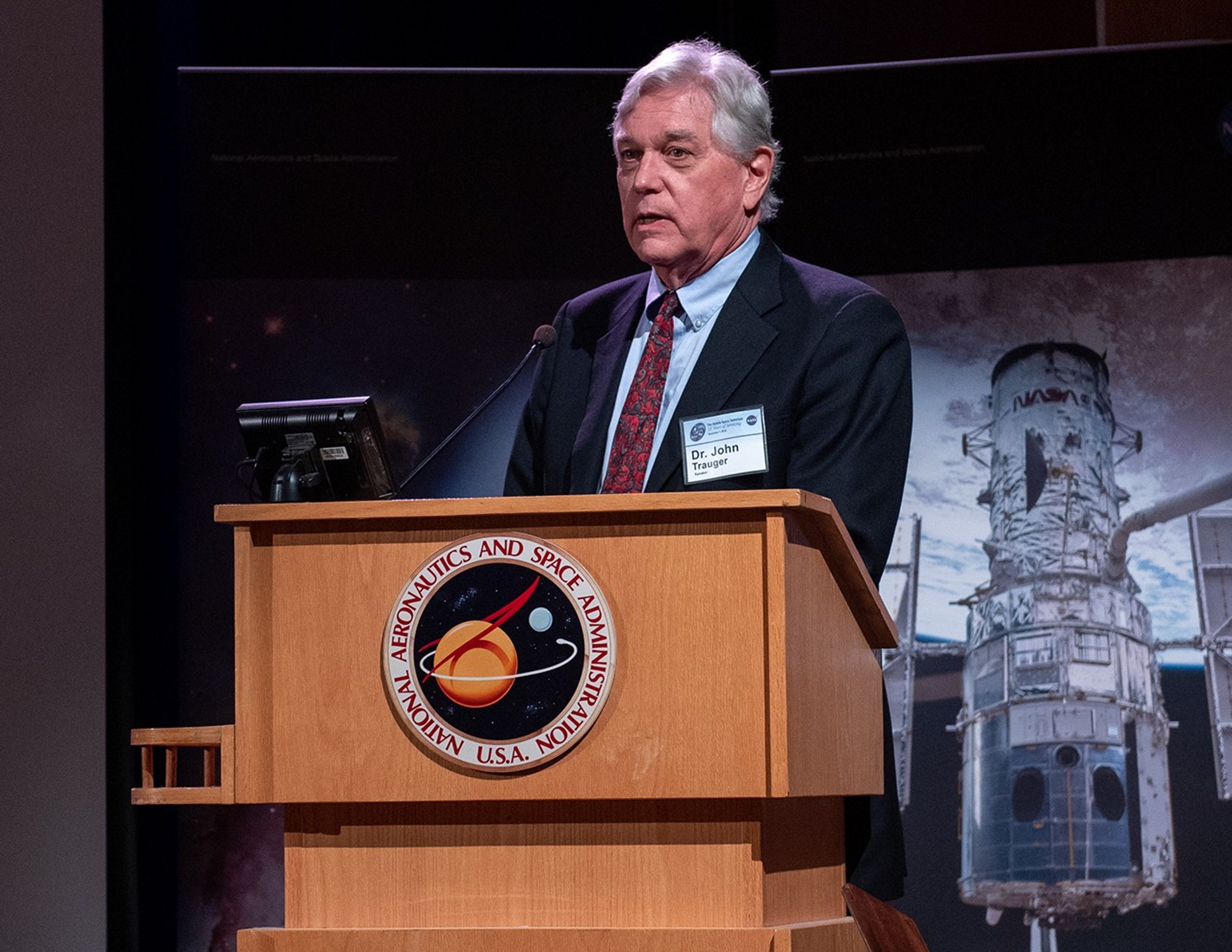 John Trauger stands behind the NASA podium, addressing the audience.
