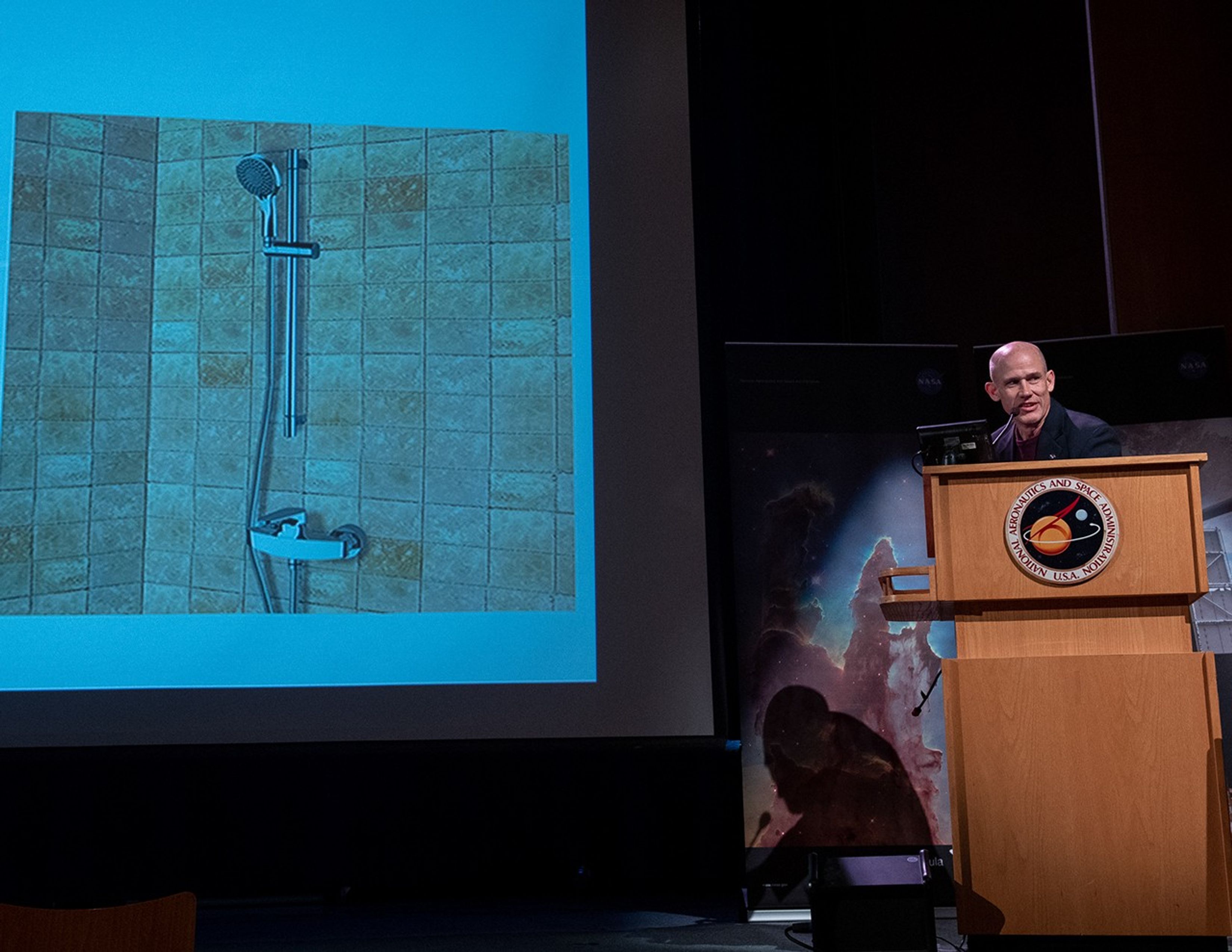 Paul Geithner stands at the podium on the right side of the image, addressing the audience. To his right, on the left, the slide show displays an image of a shower head in a bathtub/shower.