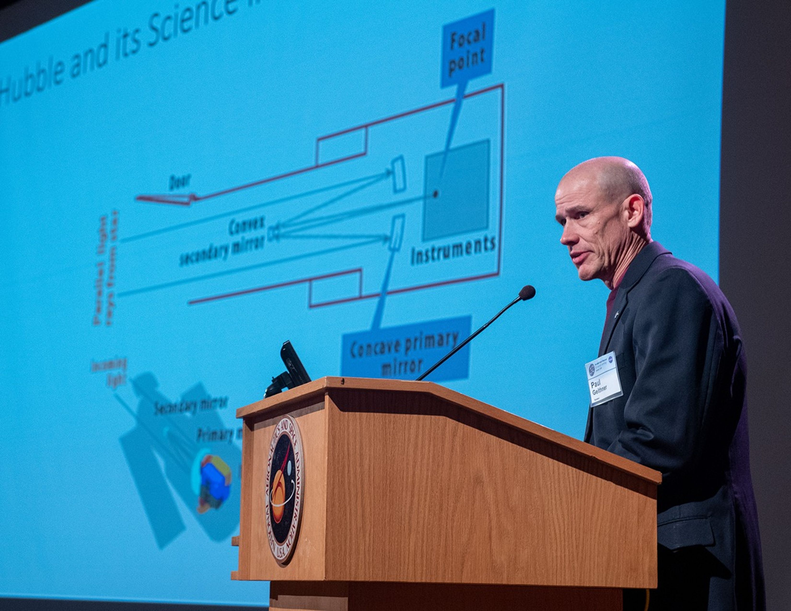 Paul Geithner addresses the audience from the podium, with the slide show--displaying a diagram of Hubble--on display behind him.