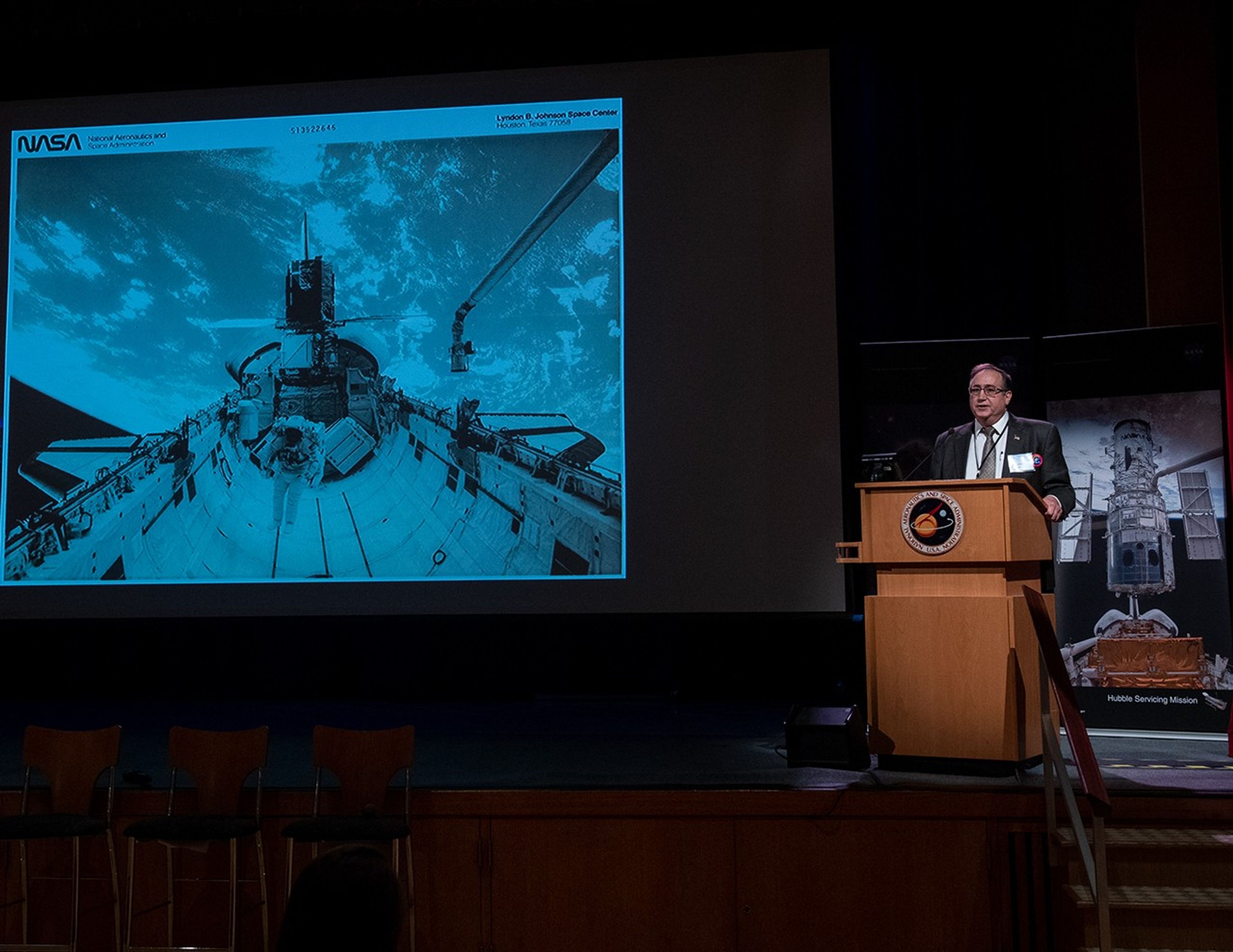 On the right side of the image, Thomas Griffin stands behind the podium, addressing the audience. On the left, the slide show displays an image taken from a servicing mission.