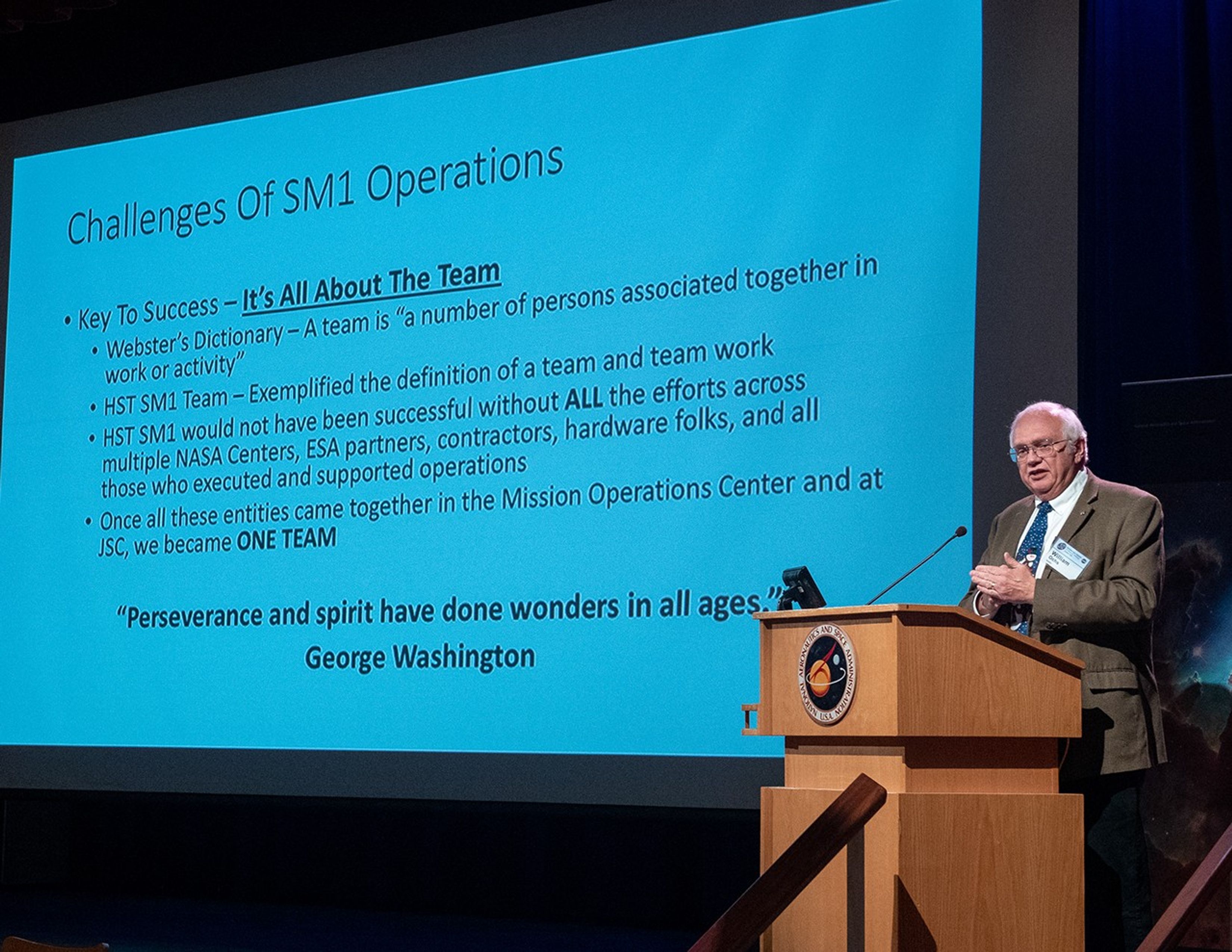 William Ochs stands at the podium, addressing the audience, on the right-hand side of the image, with the slide show on the left.