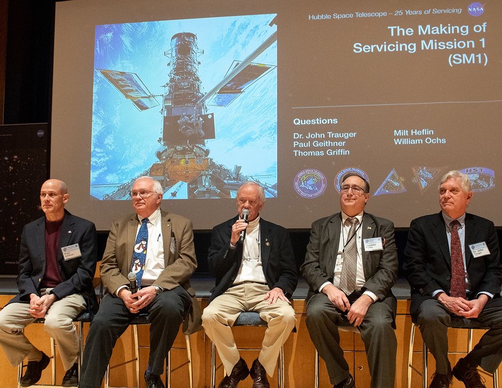 Five men sit in a row in chairs in front of the stage as part of a panel, with the slide show on the stage in the background. The man in the center, Milt Heflin, holds a microphone and speaks to the audience.