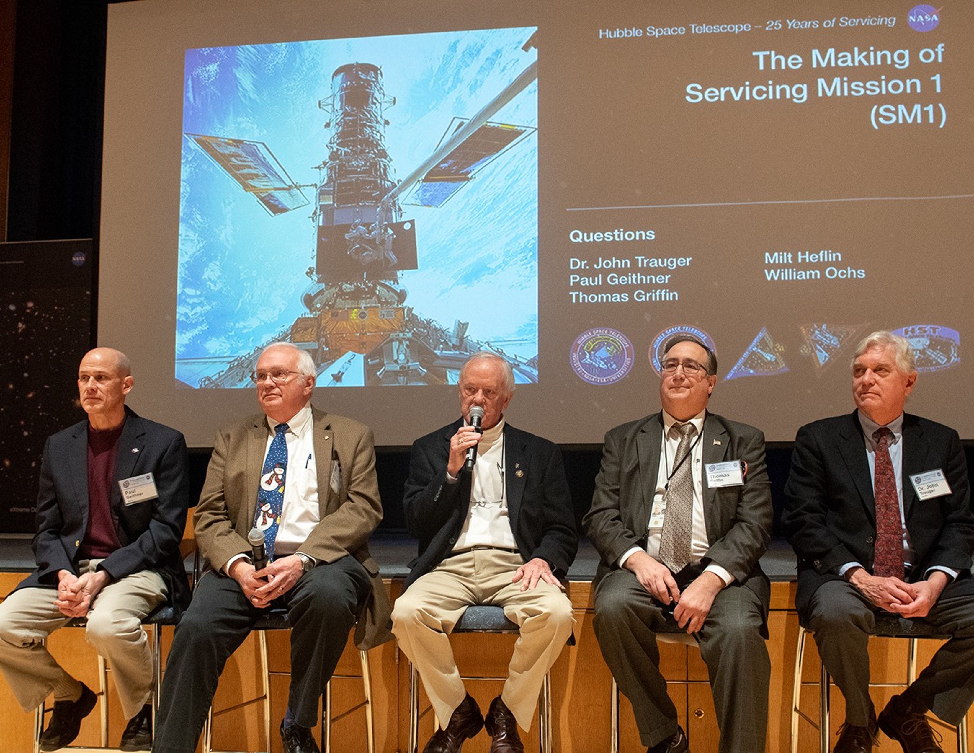 Five men sit in a row in chairs in front of the stage as part of a panel, with the slide show on the stage in the background. The man in the center, Milt Heflin, holds a microphone and speaks to the audience.