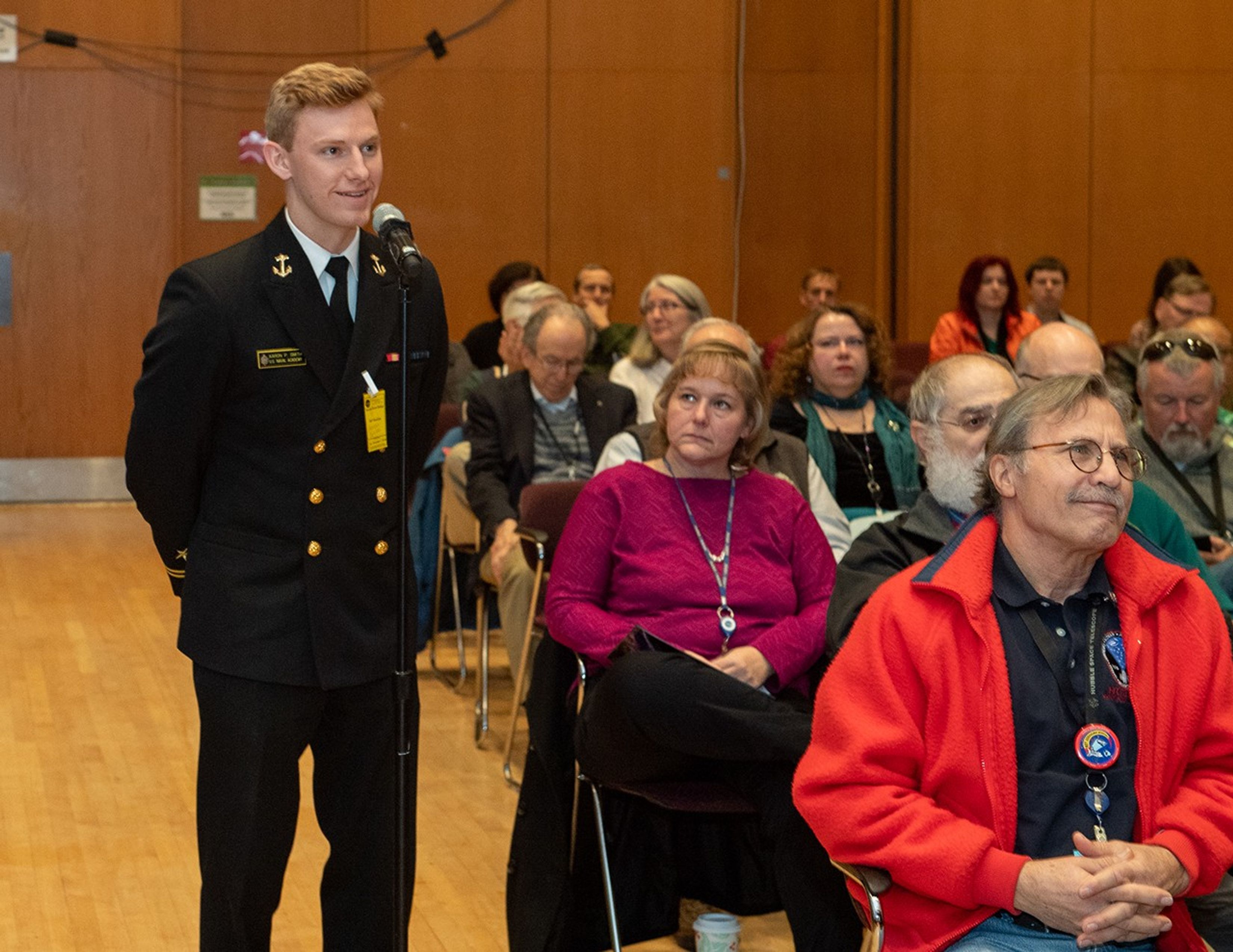 A young man in a Naval Academy uniform speaks into the audience microphone on the left-hand side of the image. The audience can be seen on the right, many of whom are glancing at the young man speaking.