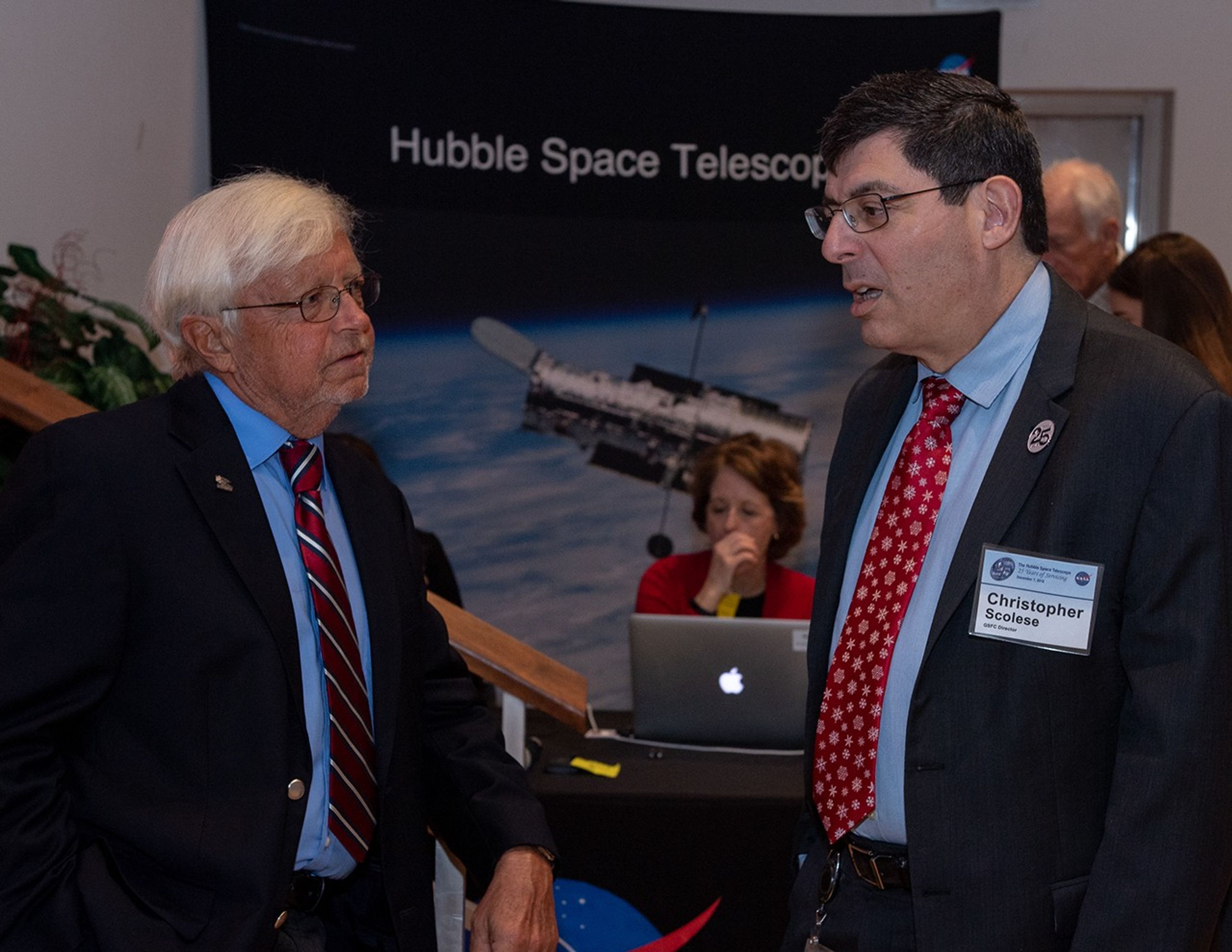 Two men talk while, in the background, a woman on a laptop sits at a greeting table with two others standing on the right.