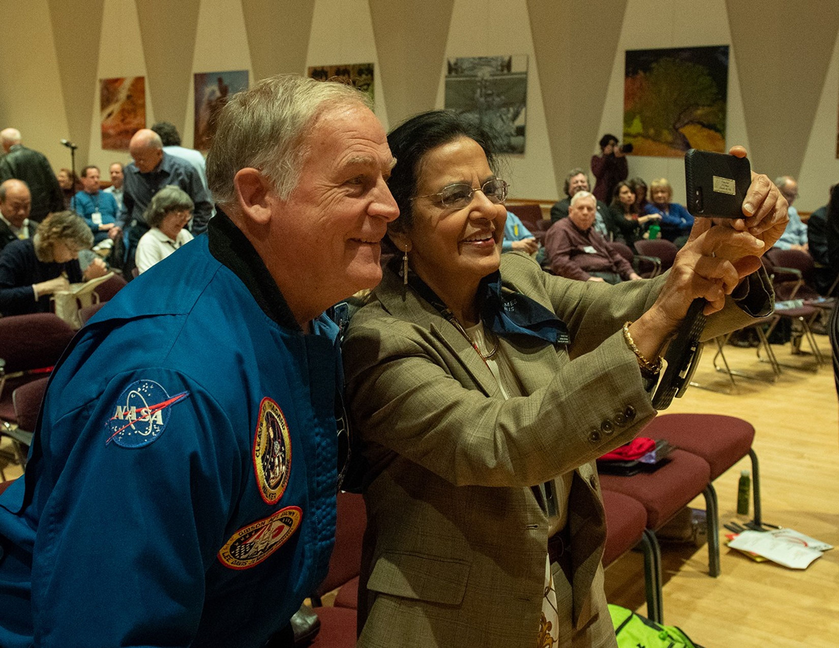 A man (astronaut) and woman (scientist) take a selfie together, with many other audience members visible in the background.