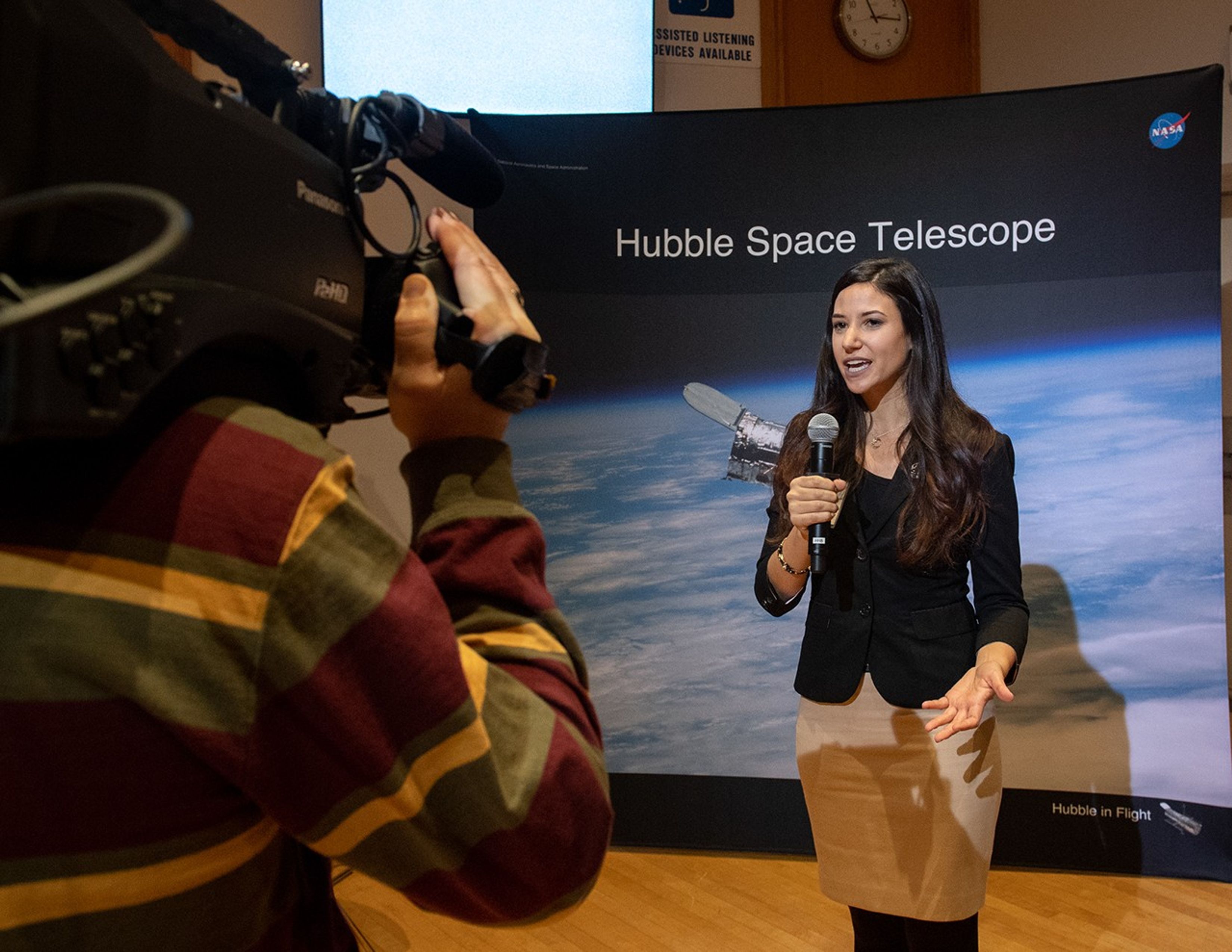 A woman, Erin Kisliuk, speaks into a microphone with an image of Hubble in the background, while a man with his back to the camera, Rob Andreoli, films her.