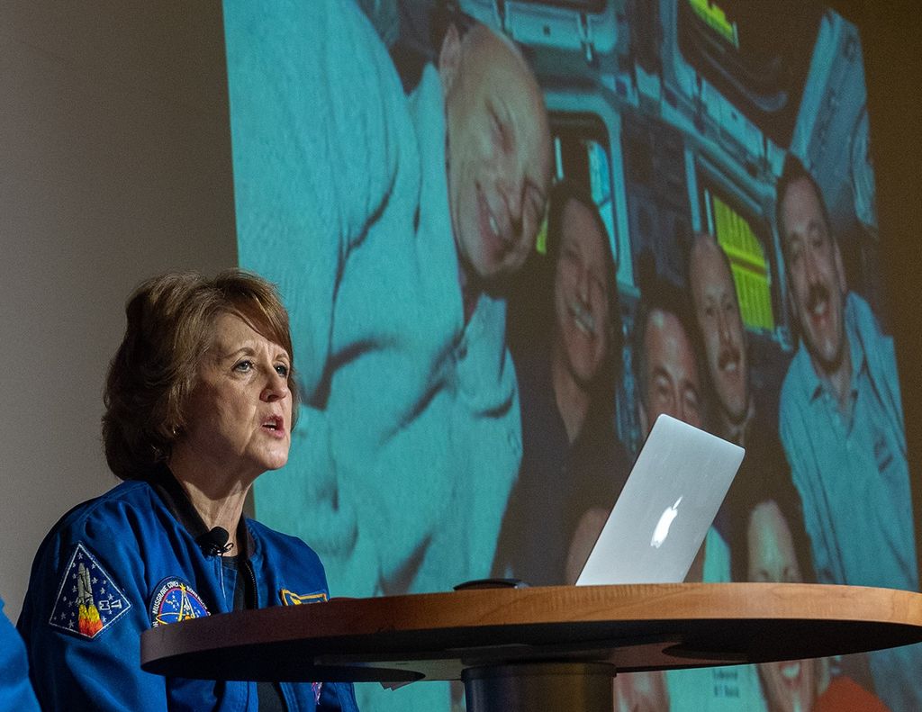 Kathy Thornton sits at a table on stage with her laptop, addressing the audience, with a picture of Hubble astronauts in the background.