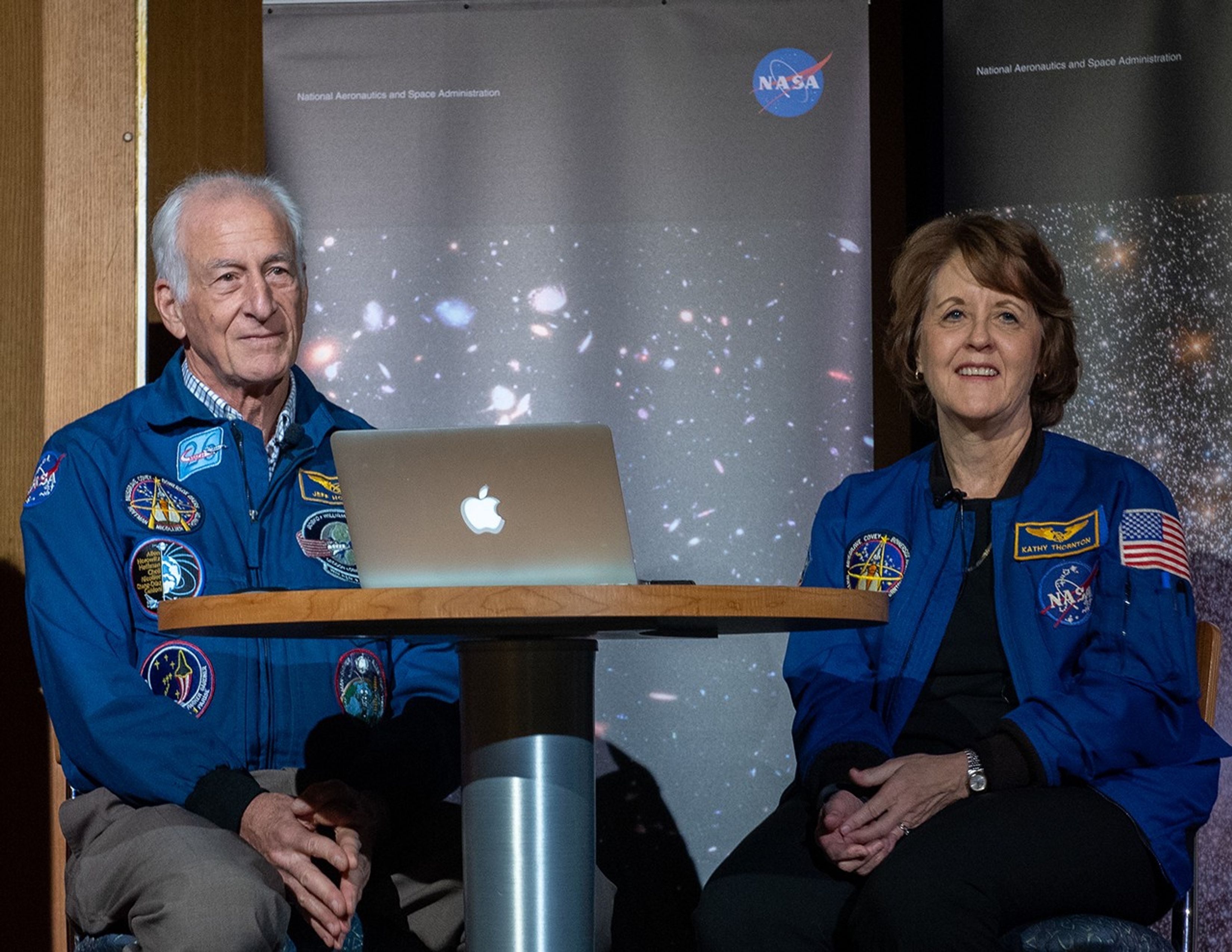 Two astronauts, Jeffrey Hoffman (left) and Kathy Thornton (right),  sit at a table onstage with a laptop on the table and Hubble images in the background as banners.