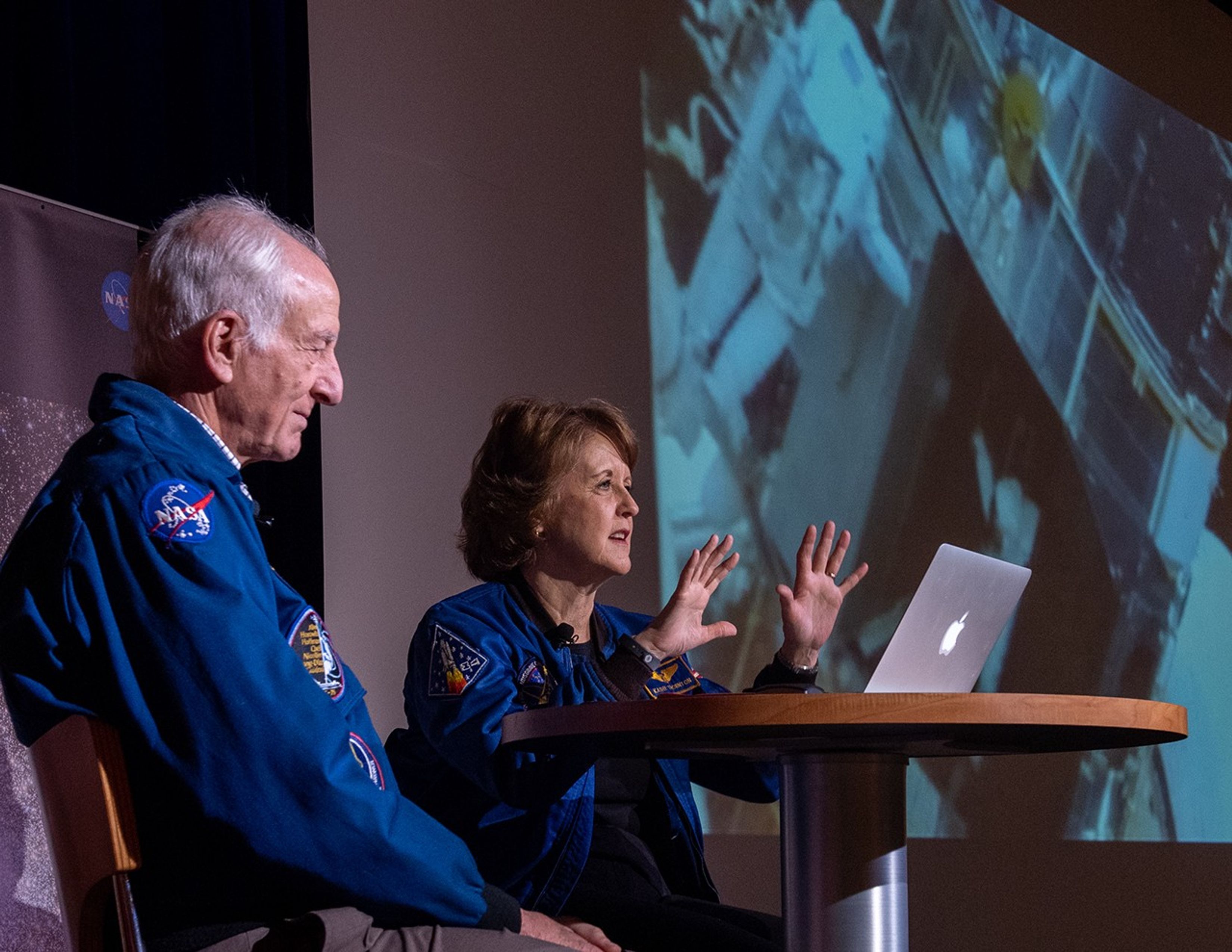 Two astronauts, Jeffrey Hoffman (left) and Kathy Thornton (right), sit at a table onstage with a laptop on the table, as Kathy speaks to the crowd.