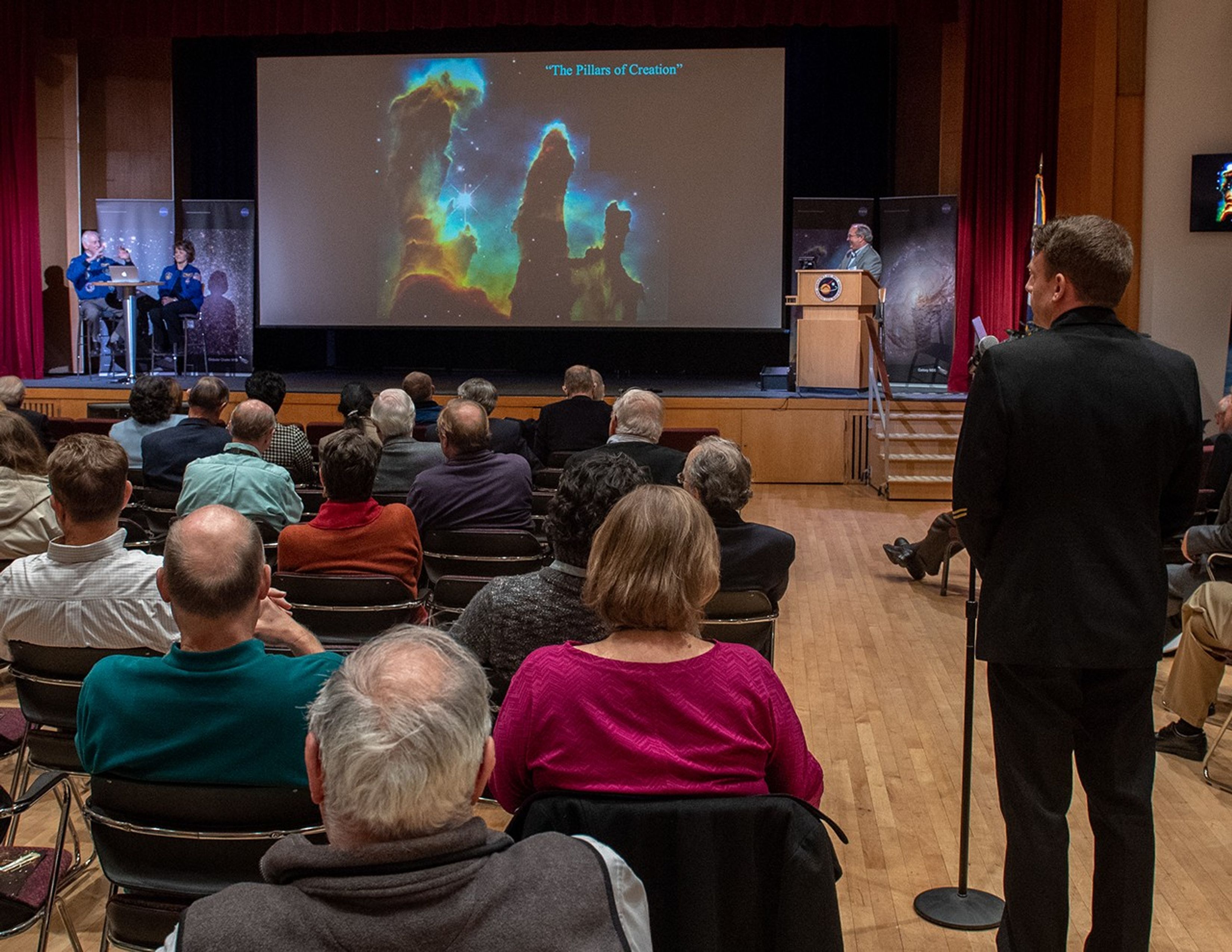 An audience member stands at the microphone on the right to ask a question; he and the rest of the audience face away from the camera. Onstage, an image of the Pillars of Creation is shown on the screen, with Ken Carpenter behind the podium on the right while astronauts Jeffrey Hoffman and Kathy Thornton sit at the table on the left.