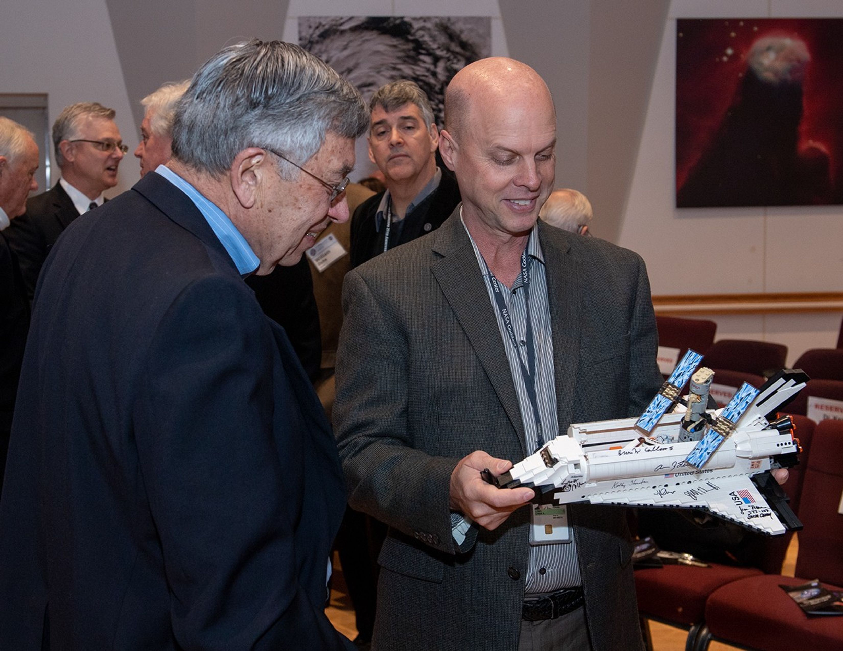 Two men admire the model of the Discovery Space Shuttle (which the man on the right is holding) with the Hubble model in it. In the background, other attendees converse, with one looking over at the model as well.