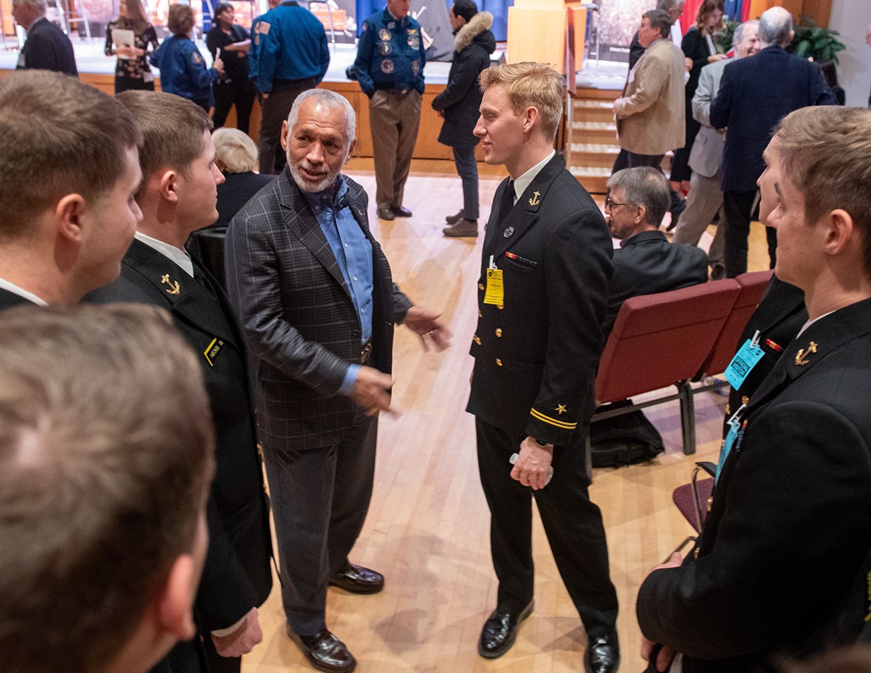 Charlie Bolden speaks with a group of six Navy midshipmen, with numerous other audience members and panelists mingling in the background.