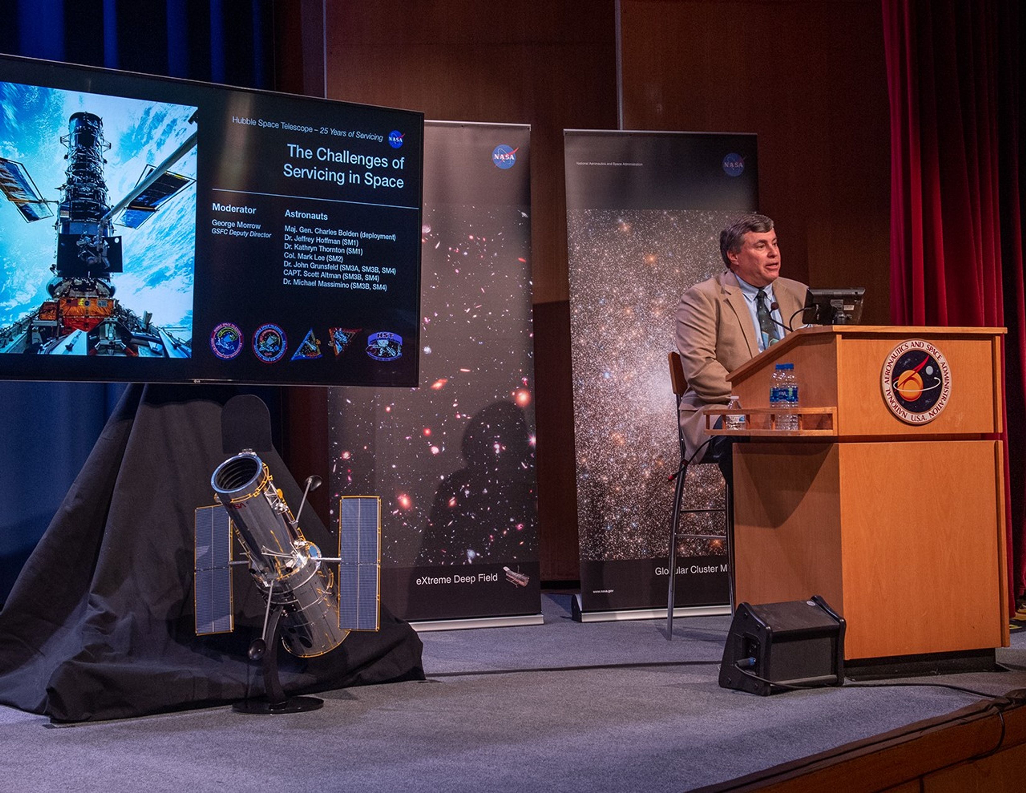 George Morrow sits at the podium, addressing the audience, on the right side of the image. On the left side, a scale model of Hubble sits on the stage beneath the slide presentation.
