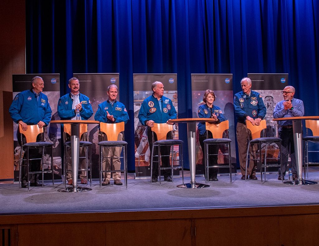Seven astronauts stand in a row behind their chairs on the stage, with five Hubble banners in the background.