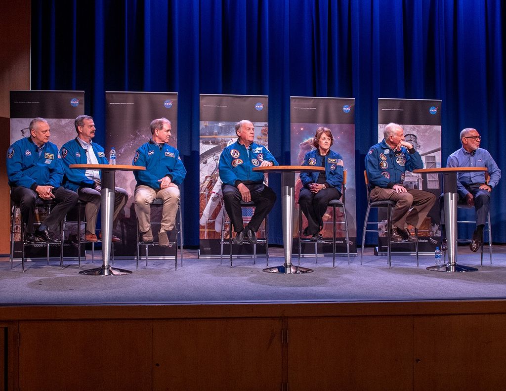 Seven astronauts sit in a row in chairs on the stage, with five Hubble banners in the background.