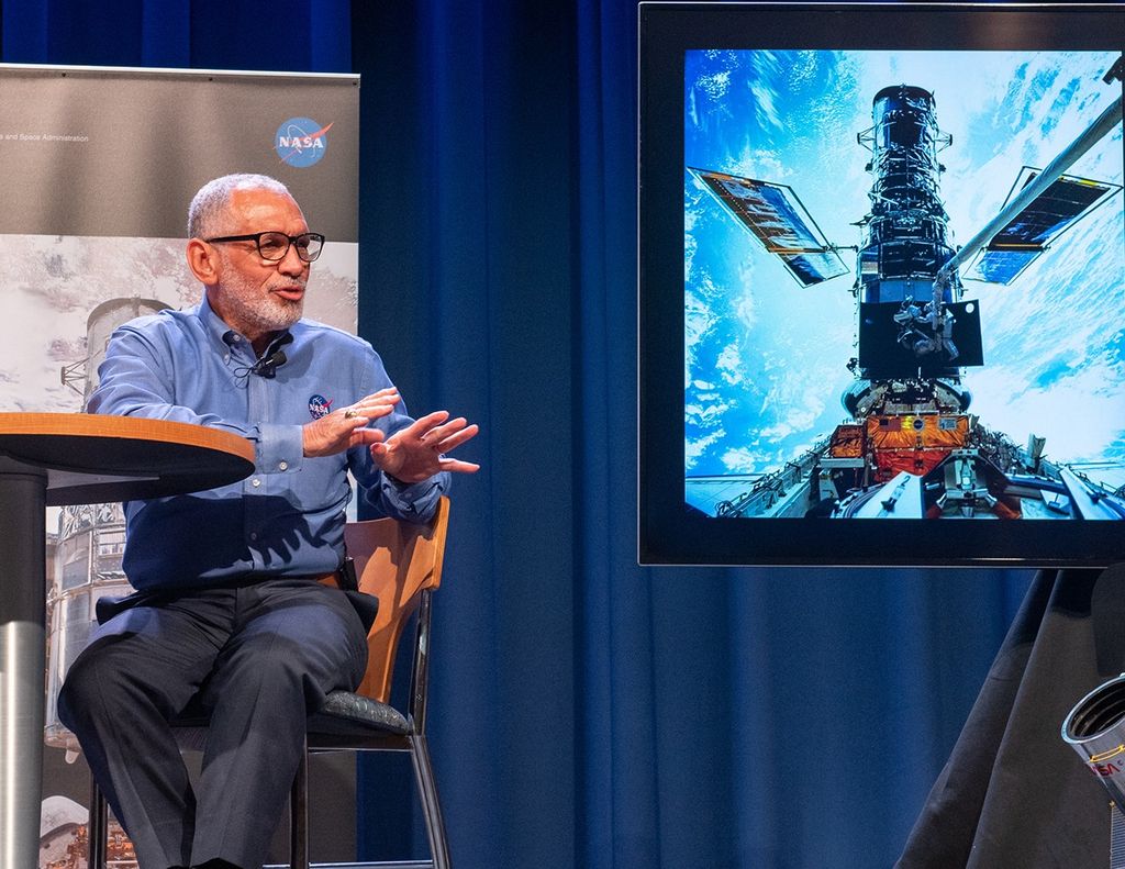 Charlie Bolden sits in a chair at a table onstage on the left side of the image. On the right side, a screen displays an image from the first Hubble servicing mission.