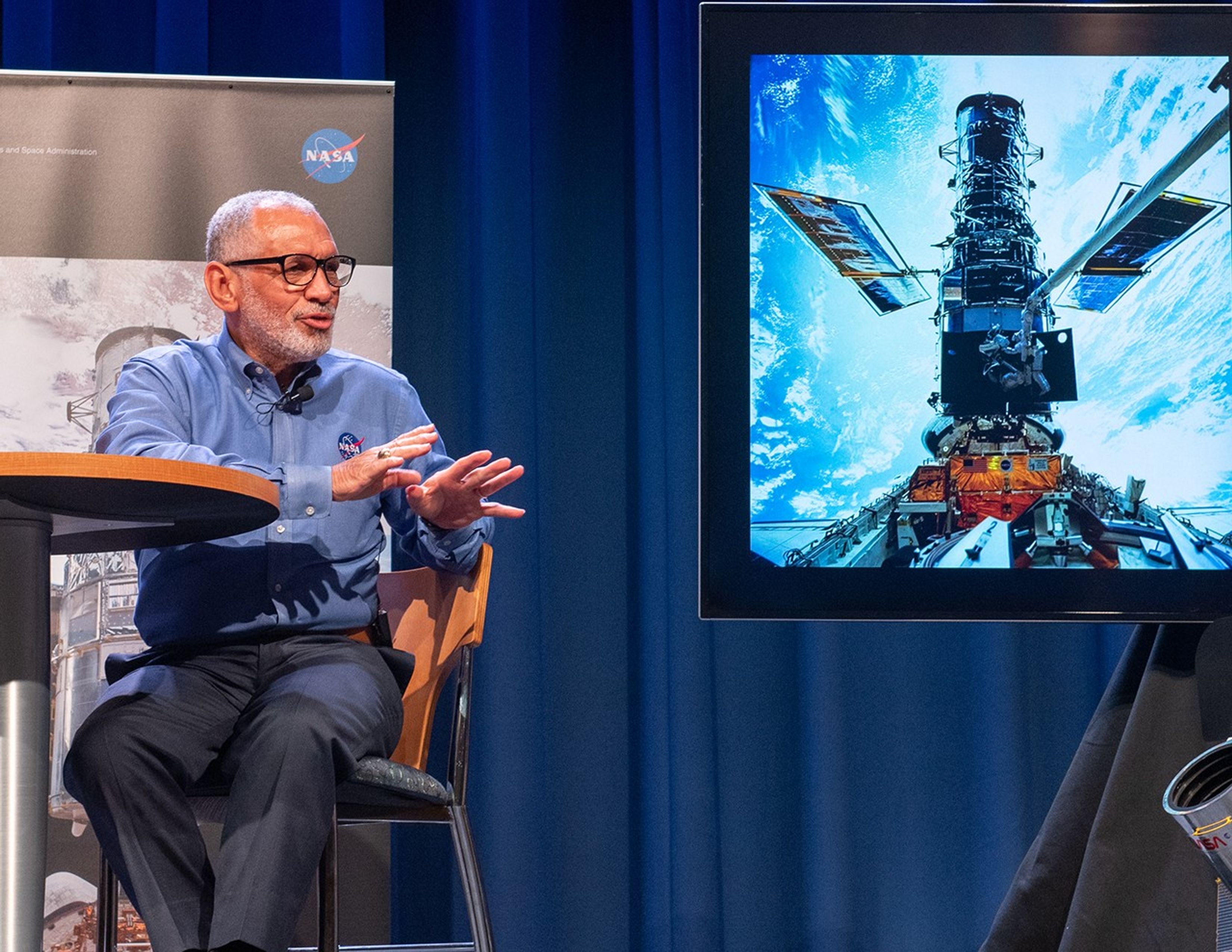 Charlie Bolden sits in a chair at a table onstage on the left side of the image. On the right side, a screen displays an image from the first Hubble servicing mission.