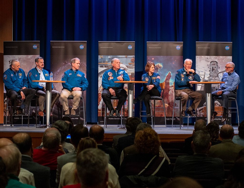 Seven astronauts sit in a row in chairs on the stage, with the other six looking toward Jeffrey Hoffman (second from the right) as he speaks. Five Hubble banners are visible in the background.