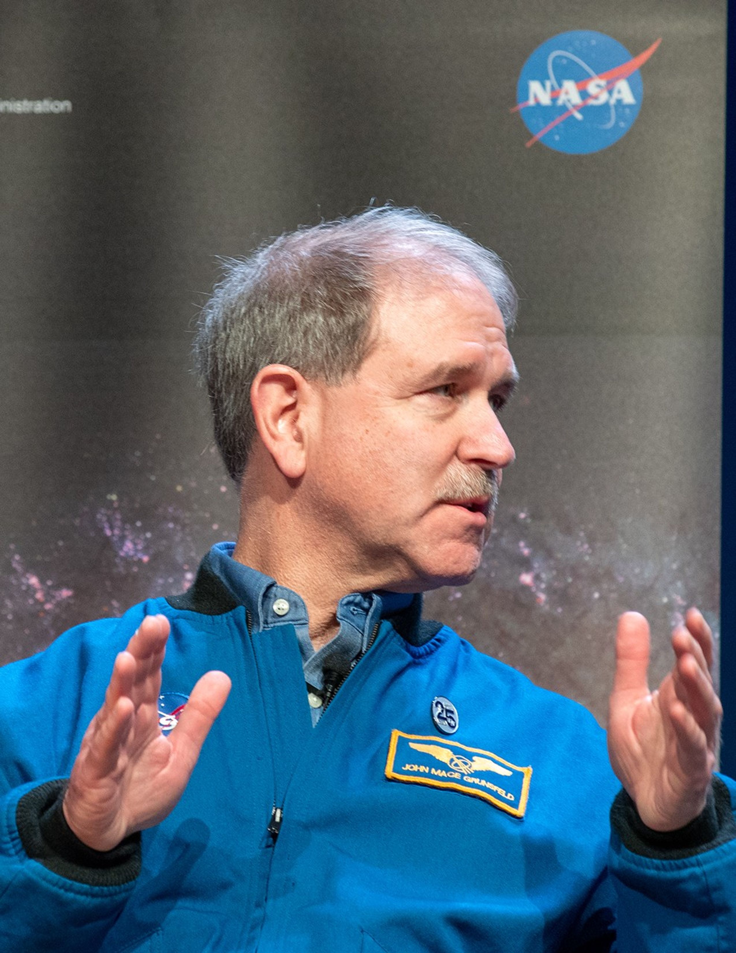 John Grunsfeld speaks to the audience while looking toward the right side of the image.