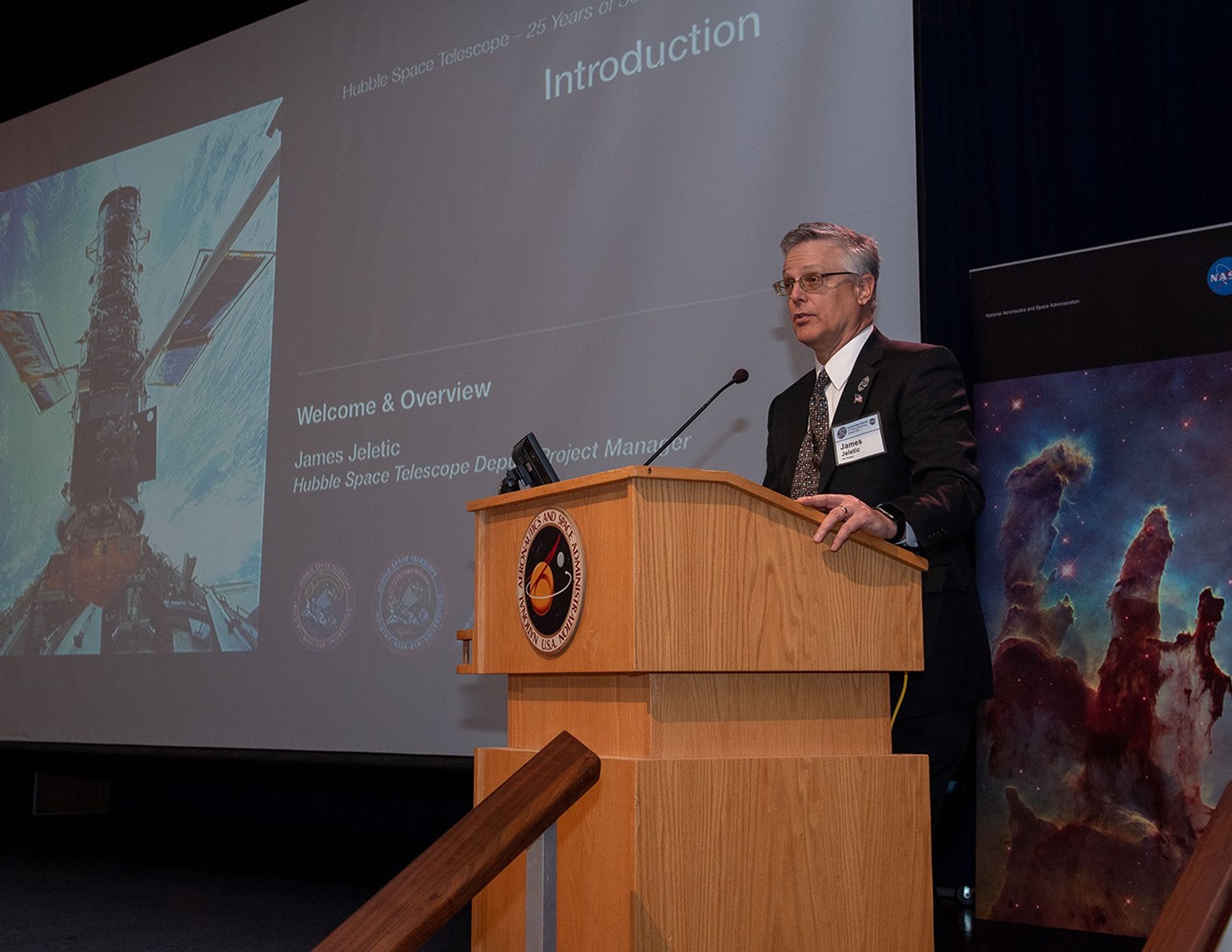 Jim Jeletic stands at the NASA podium, addressing the audience. Behind him on the right is a banner of the Pillars of Creation, and behind him on the left is the screen on which a PowerPoint presentation is being projected.
