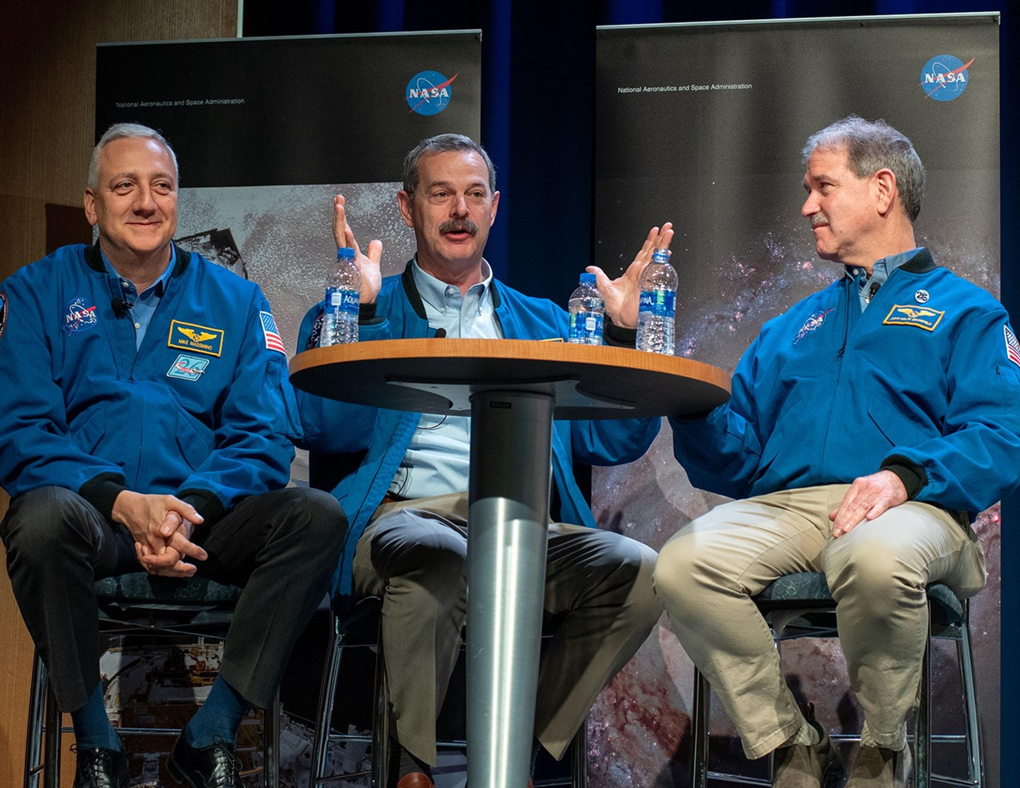 Scott Altman, center, sits at a table onstage while addressing the audience, while Mike Massimino (left) and John Grunsfeld (right) listen.