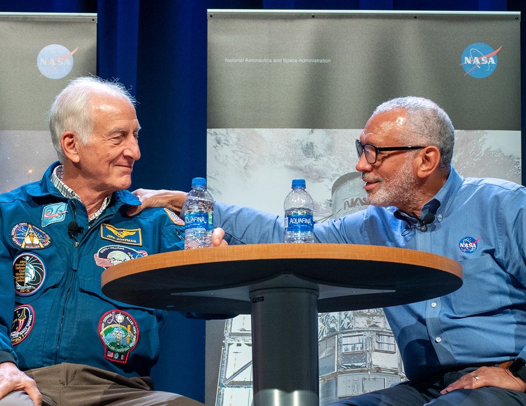 Jeff Hoffman (left) and Charlie Bolden (right) sit at a table onstage and smile at each other, with Bolden affectionately grasping Hoffman's shoulder.