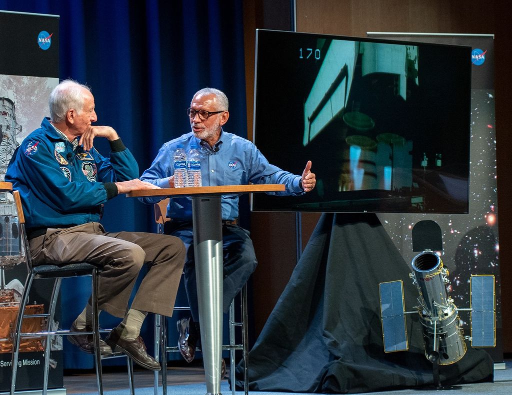 Charlie Bolden, right, talks to Jeffrey Hoffman, left, as they both sit at a table onstage on the left side of the image. On the right, an image shows footage from a previous servicing mission, while a model of Hubble sits below the screen.