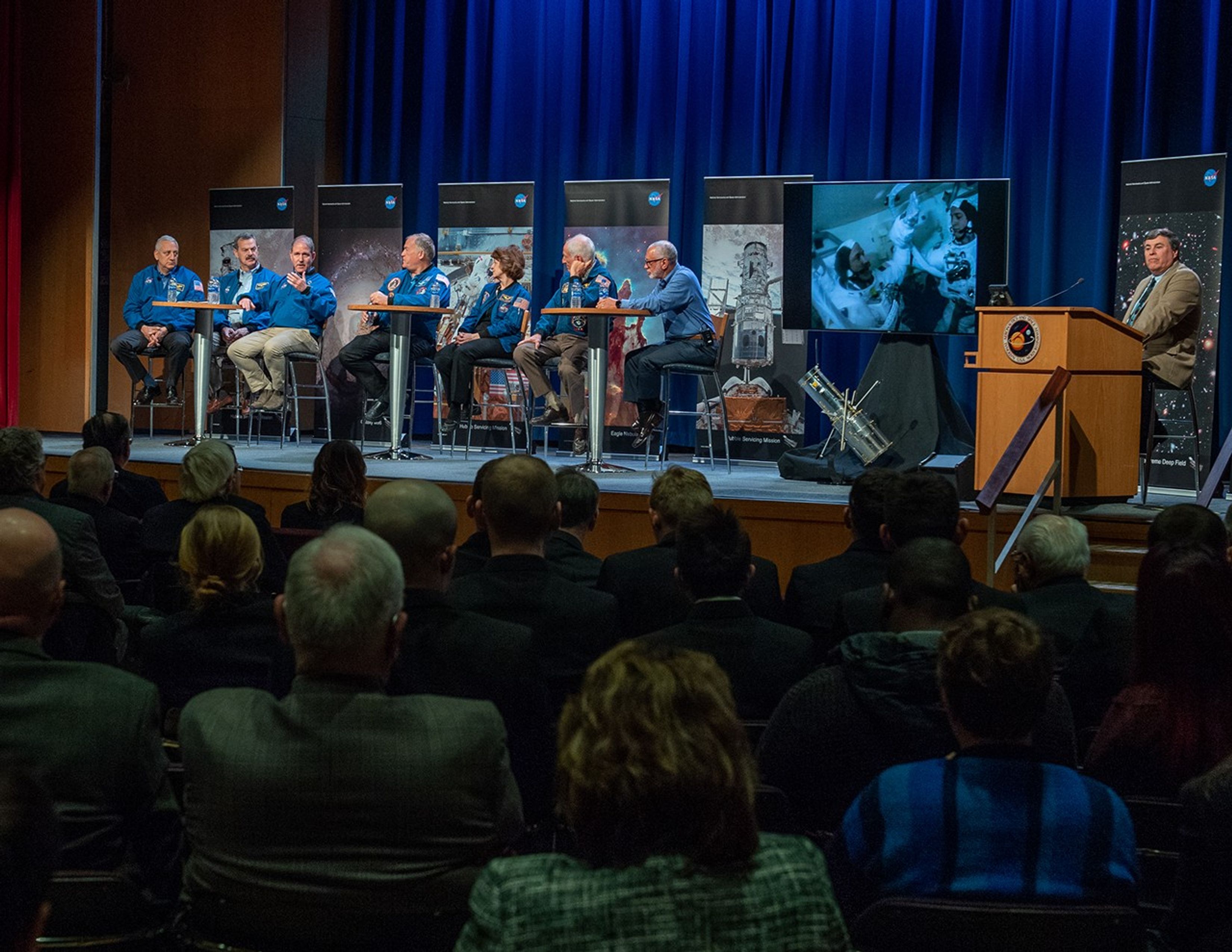 Astronaut John Grunsfeld speaks on the astronaut panel to an attentive audience while the other six astronauts face him, and George Morrow (far right) listens from the podium.