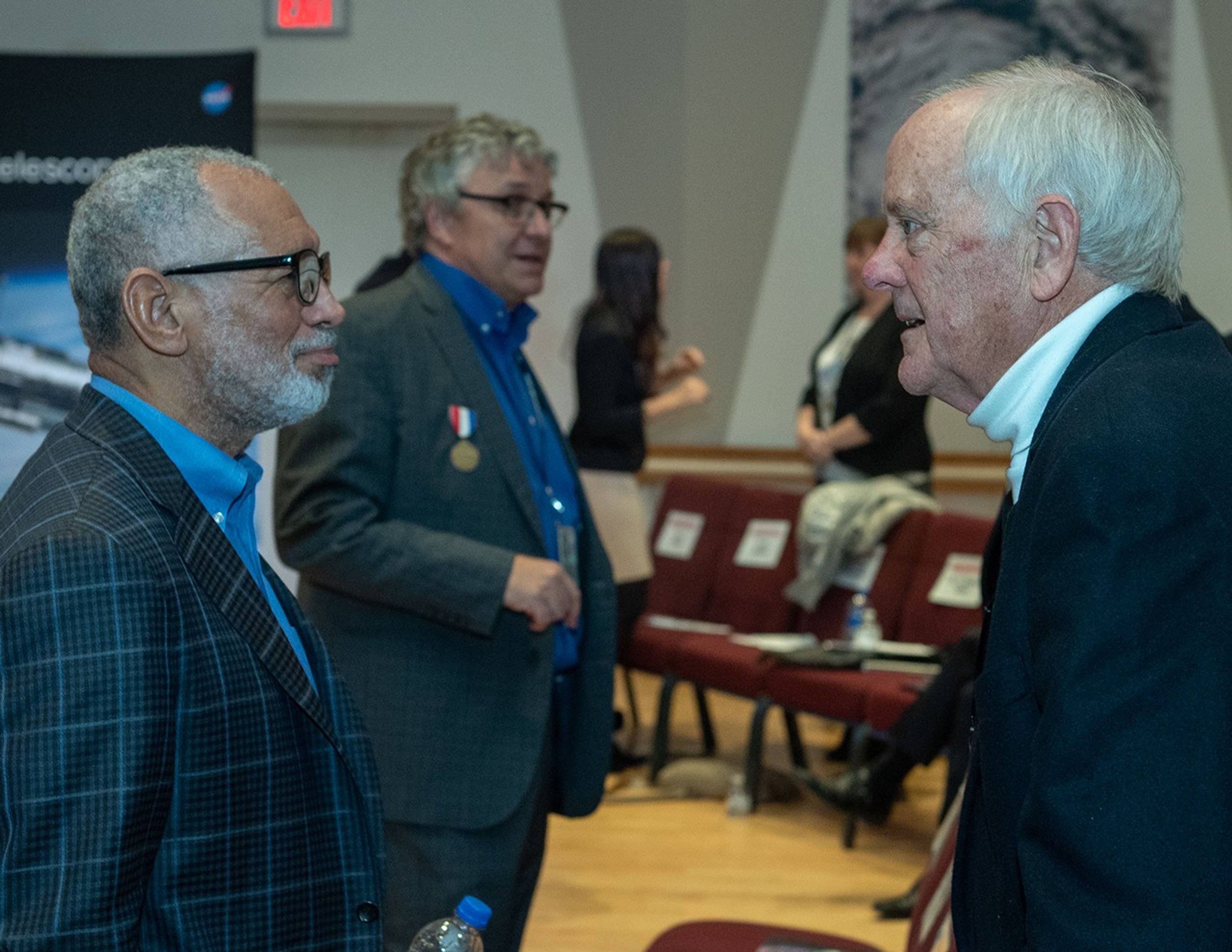 Two men, Charlie Bolden (left) and Milt Heflin (right) talk, with additional audience members visible in the background.