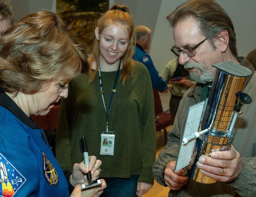 Astronaut Kathy Thornton (left) signs a solar panel from a male employee's Hubble model (right), while a female employee (center) watches in the background with a smile.