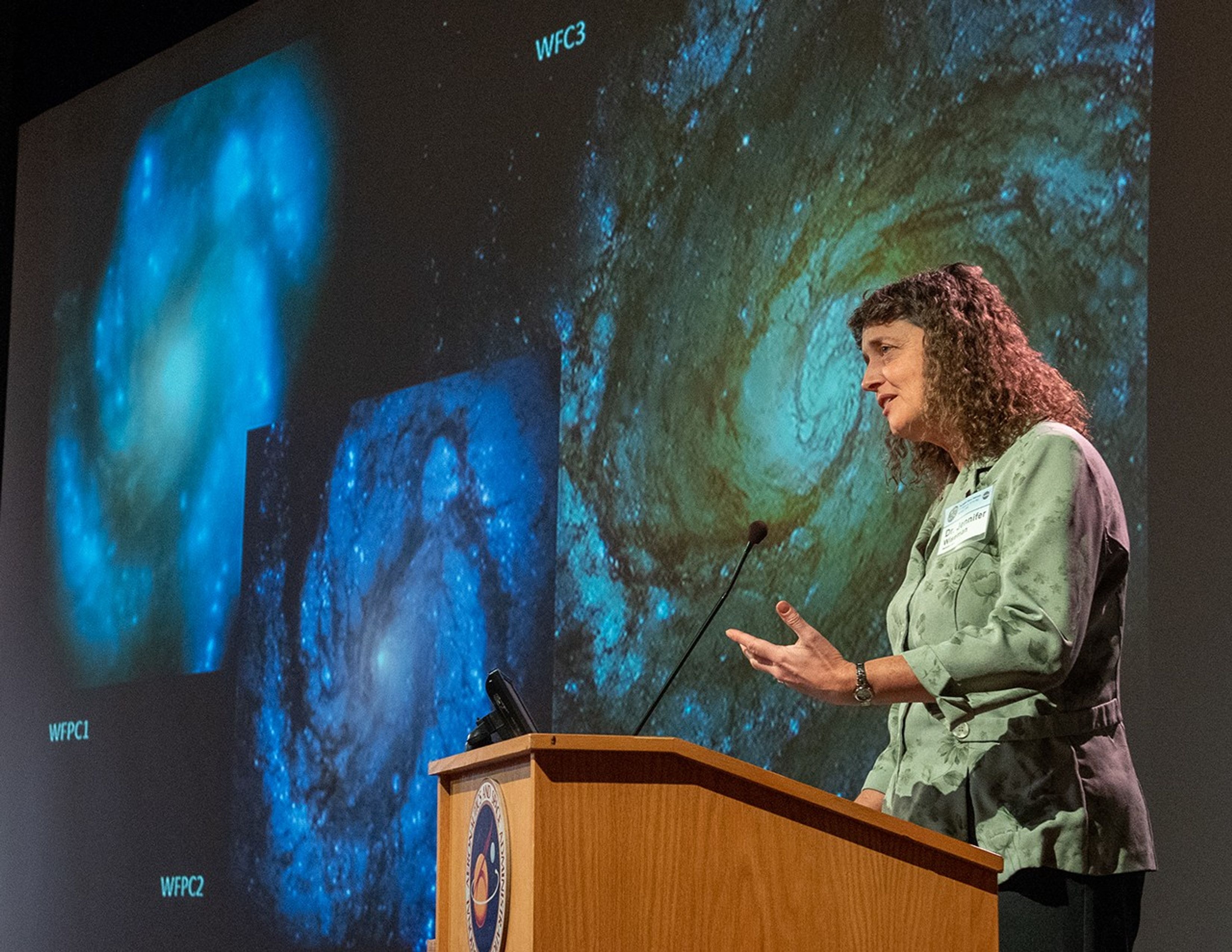 Dr. Wiseman addressing the audience from the podium, with three Hubble images of the same galaxy (taken at different times by different cameras) visible on the screen in the background.