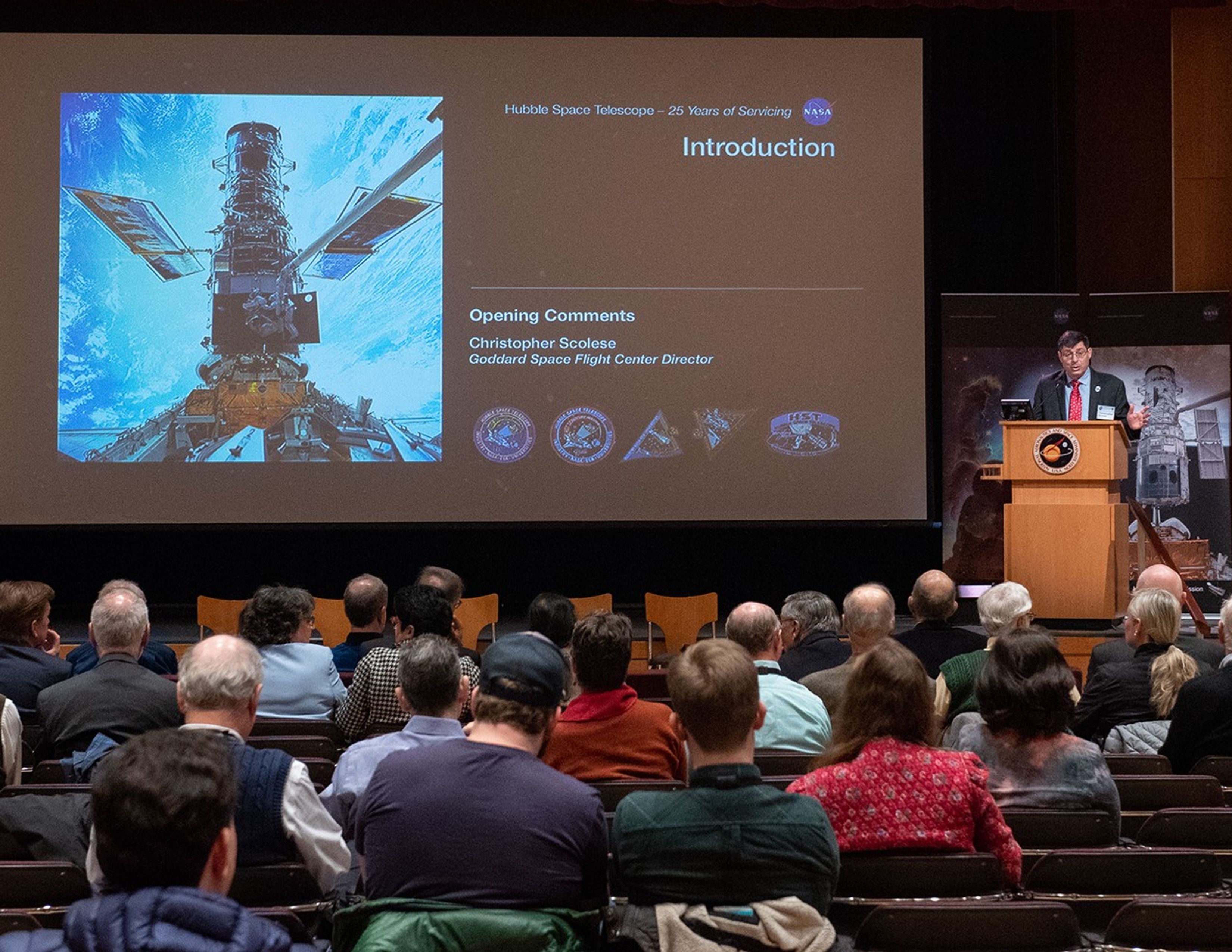 Christopher Scolese, right, addresses an attentive listening audience, while the slide show on the left shows an introduction slide with an image from a Hubble servicing mission.