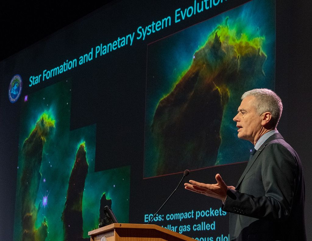 Mark Clampin speaks from the podium, with Hubble images of the Pillars of Creation on the screen in the background