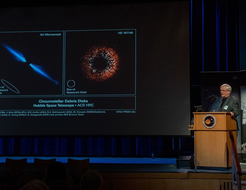 David Leckrone speaks to the audience from the podium on the right side of the image. On the left side, a Hubble science image is visible on the screen.