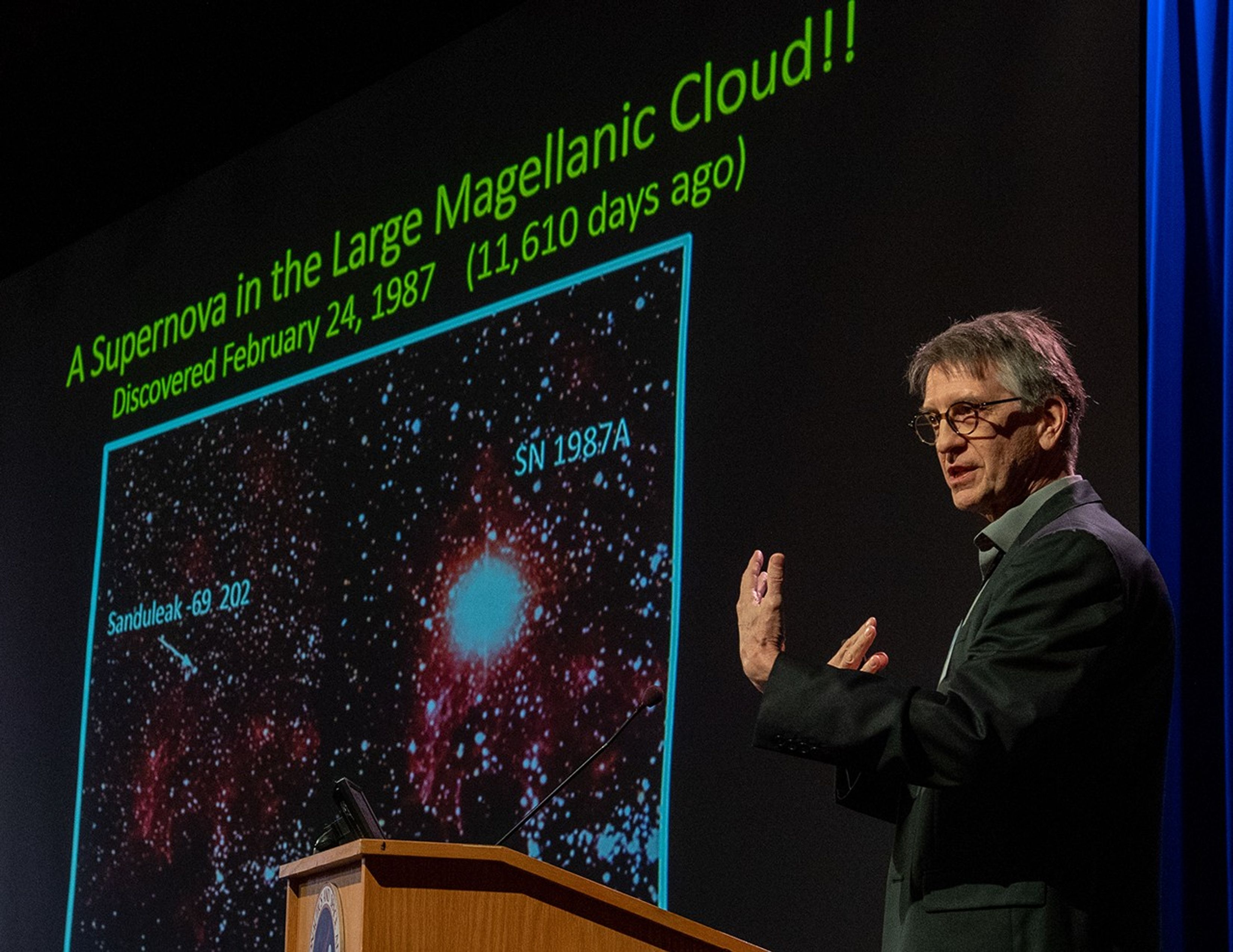 George Sonneborn speaks from the podium, with a Hubble science image of supernova 1987A visible on the screen in the background.
